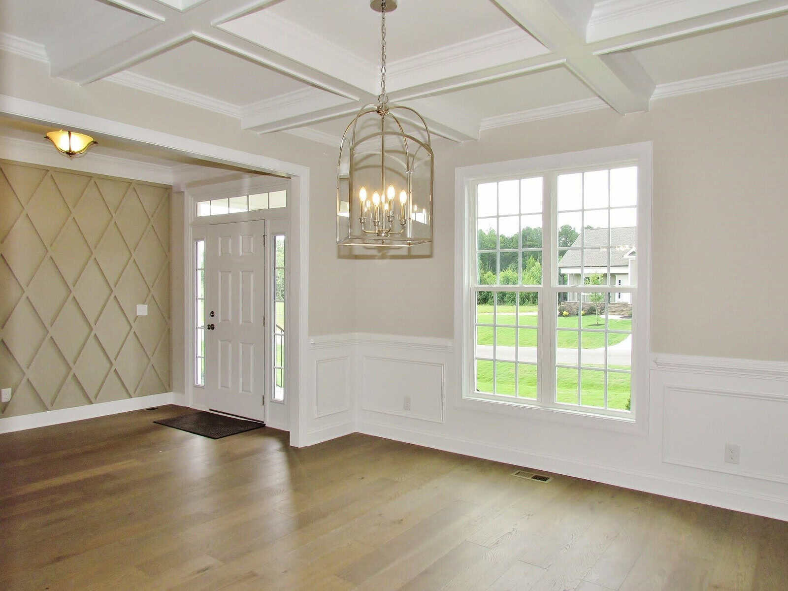 Bright, empty room with light wood floors, white wainscoting, a coffered ceiling, a large window, a modern chandelier, and a white front door with sidelights. Neutral walls, with one accent wall featuring a diamond pattern.