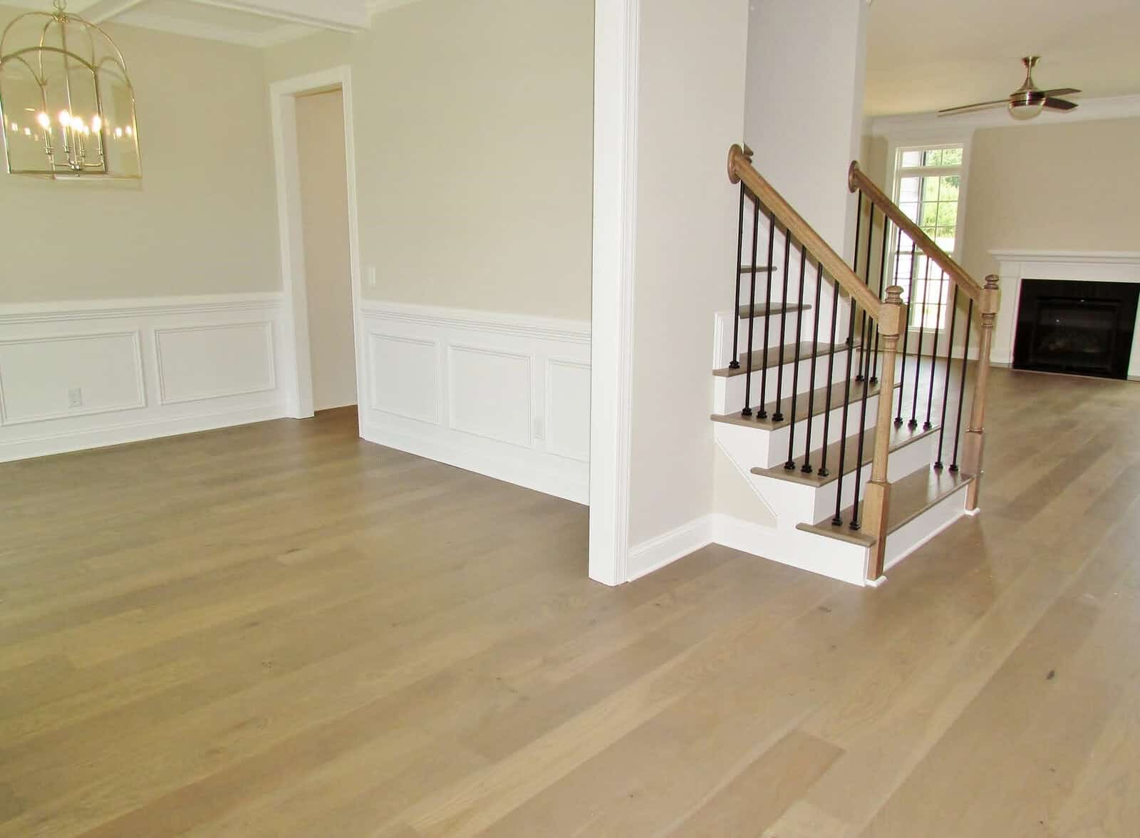 A spacious, empty living area with light wood floors, white walls, a staircase with a wooden handrail, and a gold light fixture. Large windows and a fireplace are visible in the background.
