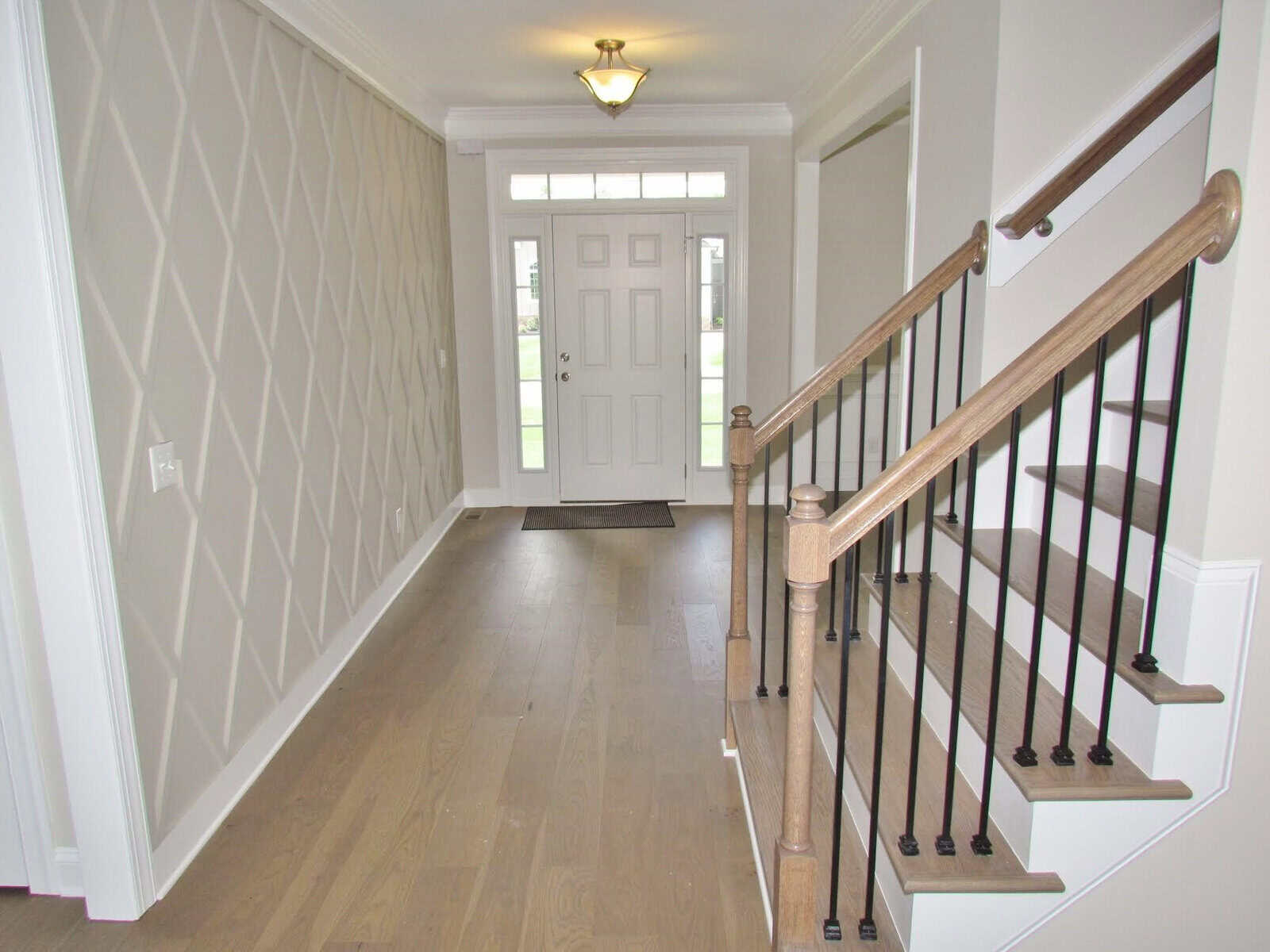 Bright entryway with light wood flooring, a patterned accent wall on the left, and a staircase with wooden handrails and black spindles on the right. White front door with windows above lets in natural light.