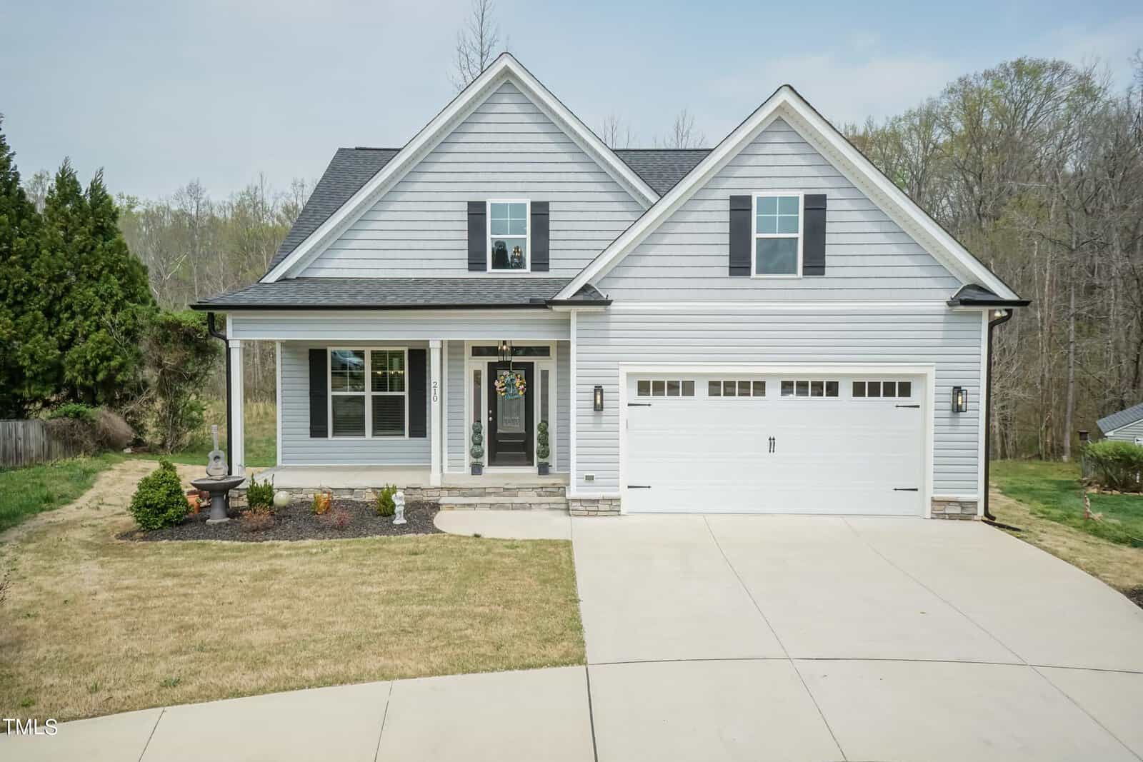 A two-story house with gray siding, white trim, and a dark roof. The front features a porch with columns, windows with shutters, and a two-car garage. The lawn is neatly landscaped with shrubs and a small tree.