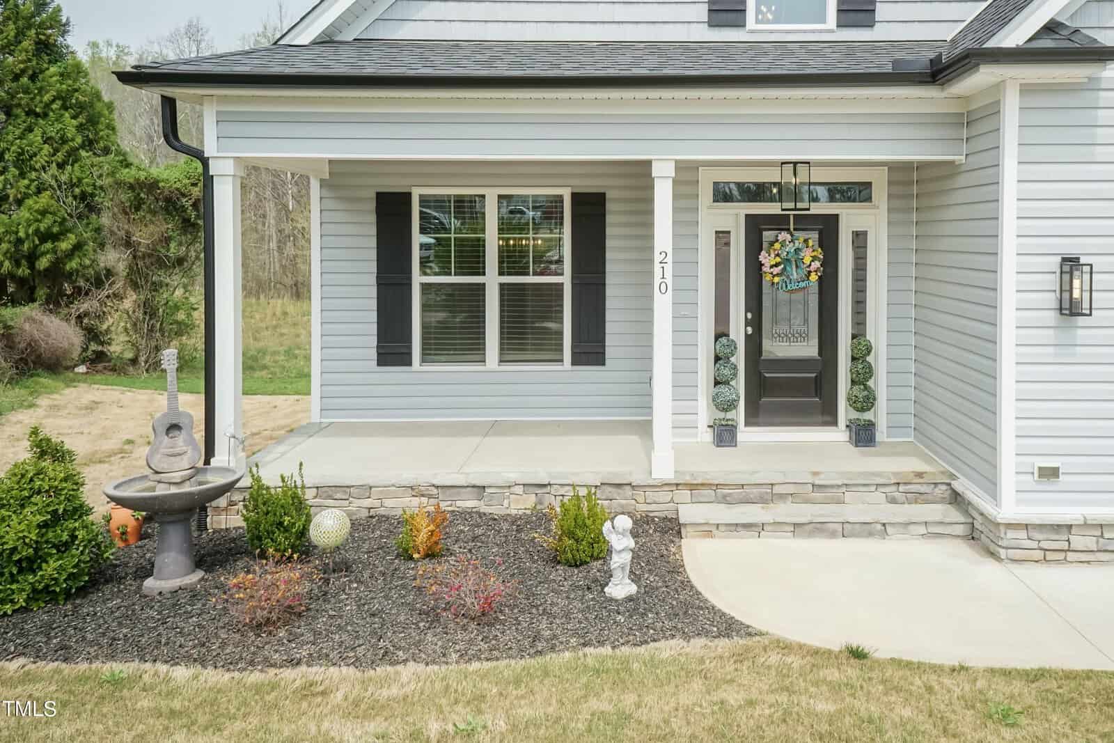 Front view of a house with light gray siding. A small garden with colorful plants and a birdbath adorned with a guitar sculpture is in the foreground. The porch features decorative topiary displays and a door with a wreath.