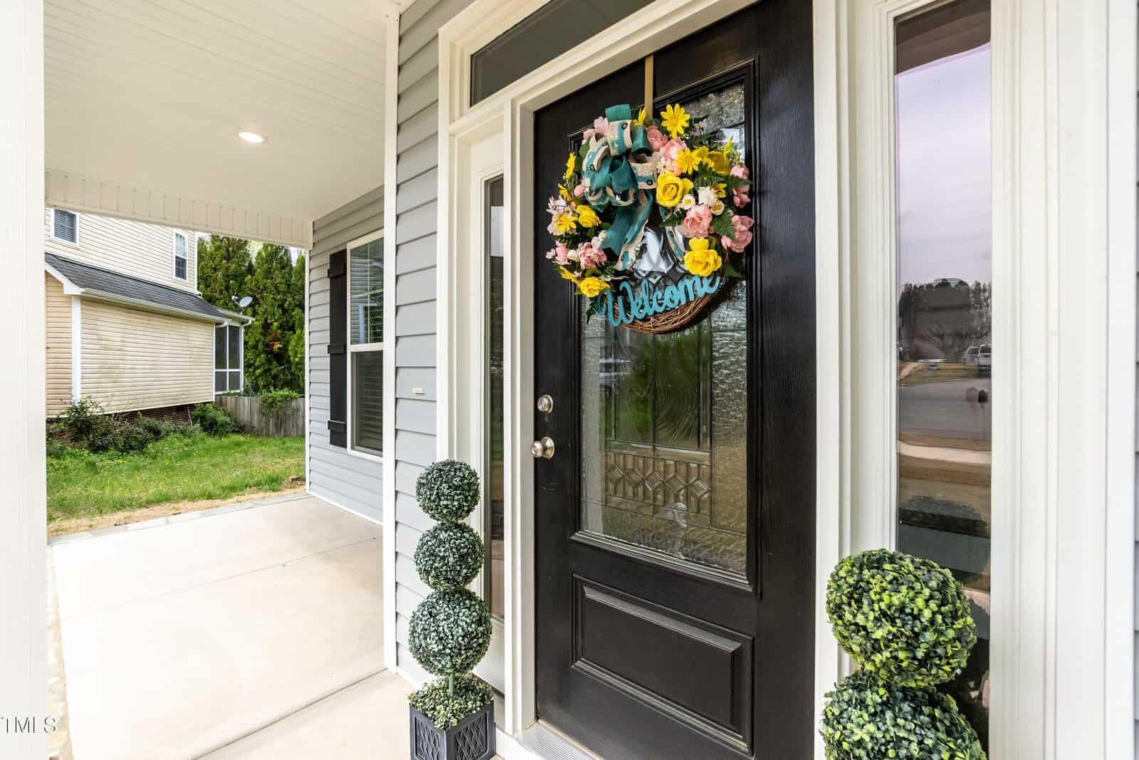 A black front door with a glass panel is decorated with a vibrant floral wreath and a "Welcome" sign. Topiary plants are placed on either side of the entrance. The house exterior is gray with white trim, and part of the driveway is visible.