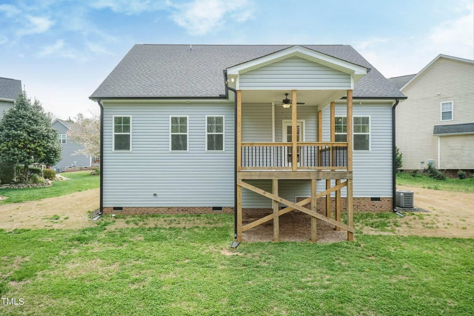 A small, light gray house with a dark roof features a wooden deck with stairs leading to the grassy backyard. The yard has patchy grass and the house has several windows and a back door.