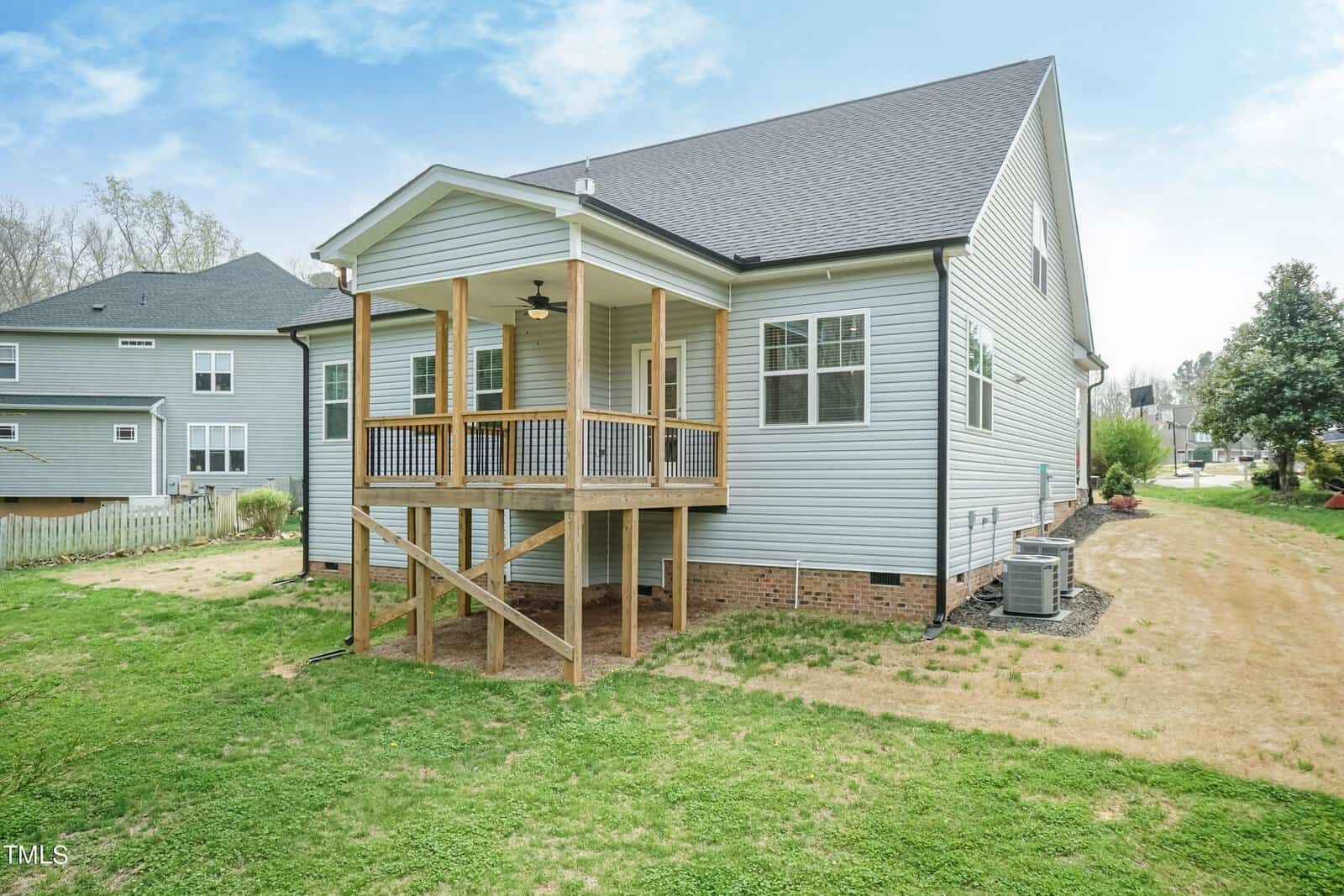 A modern gray house with a raised wooden deck and covered porch in the backyard. It features a pitched roof, a small lawn, and neighboring houses in the background under a partly cloudy sky.