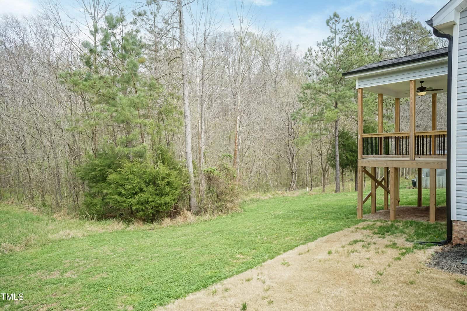 A wooden deck attached to a house overlooks a grassy backyard, bordered by a line of trees. The backyard transitions from lush green grass to dry, sparse patches near the house. A clear blue sky is visible above.
