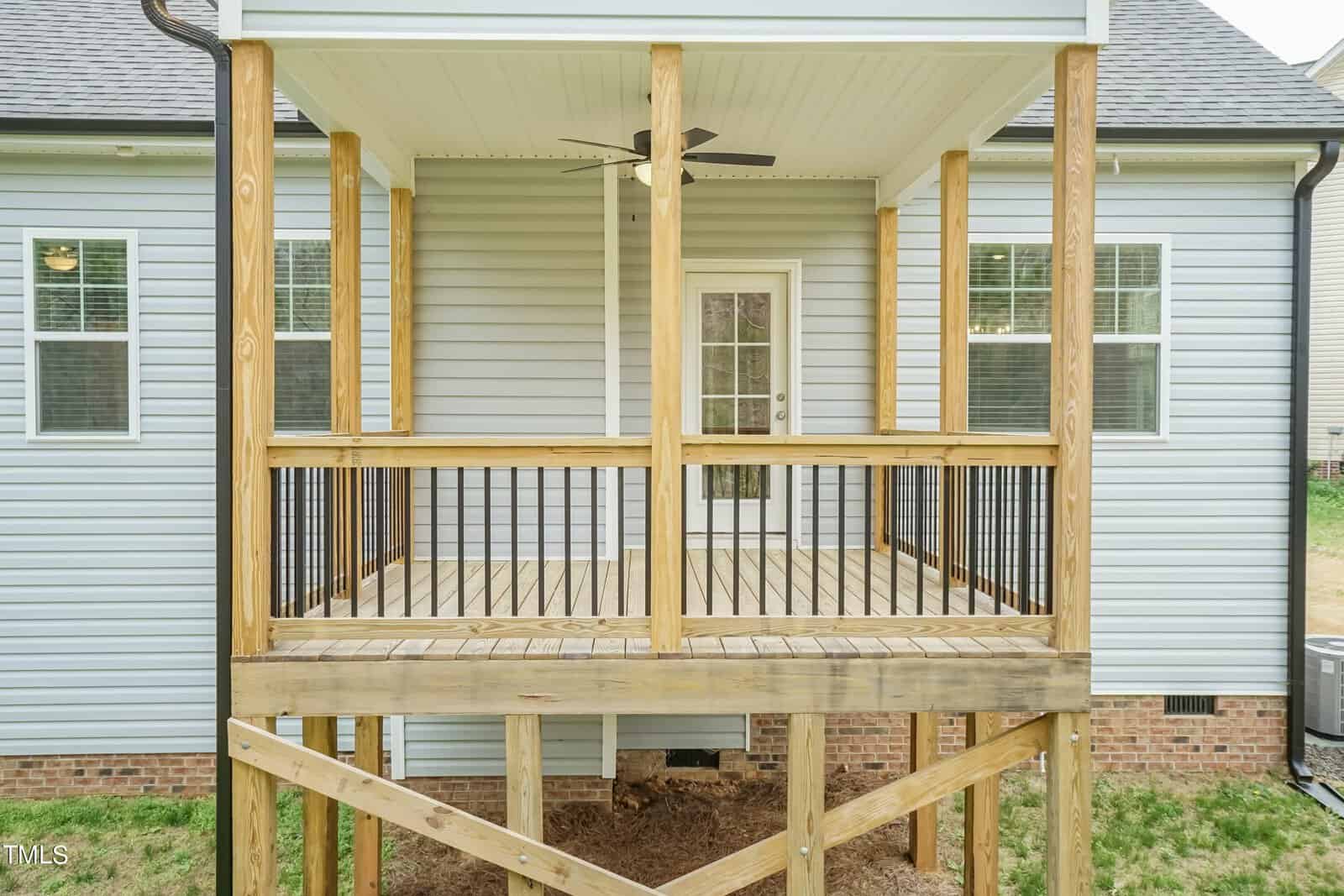 A wooden deck with railings extends from the back of a gray house with white trim. It features a glass door, two windows, and a ceiling fan. Below the deck, the base is supported by wooden posts.