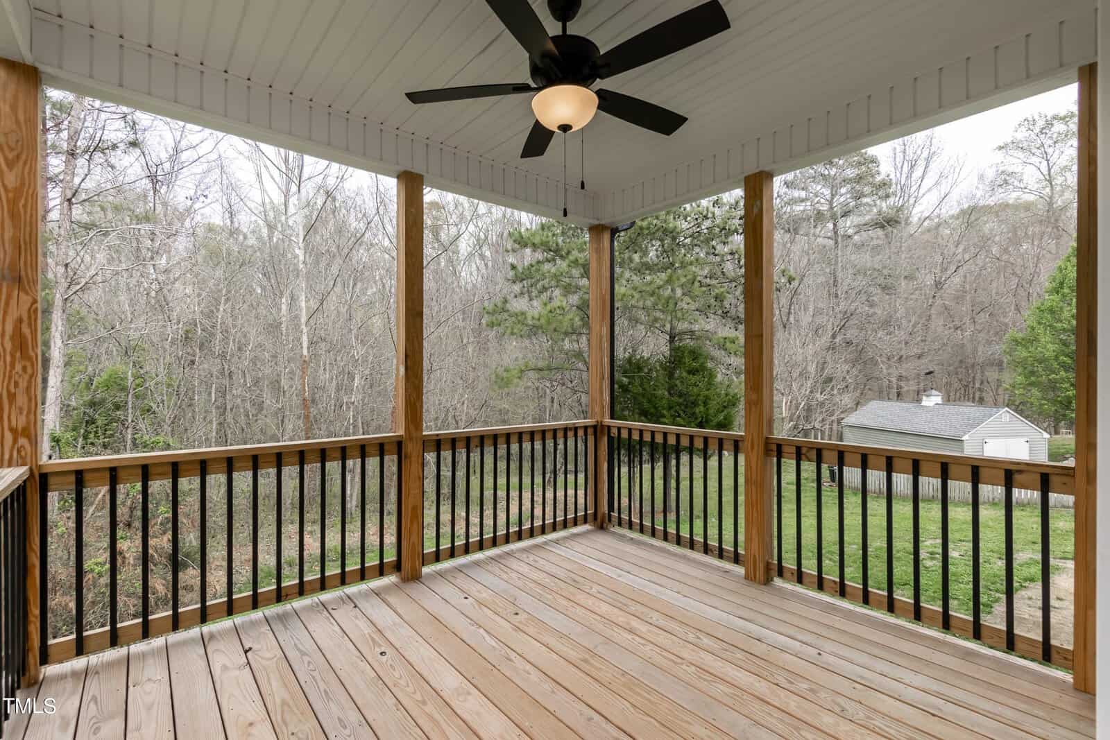 A wooden deck with a black ceiling fan and wood railing overlooks a lush, green backyard. Tall trees border the yard, and a small white shed is visible in the distance. The sky is overcast, hinting at a calm, serene setting.