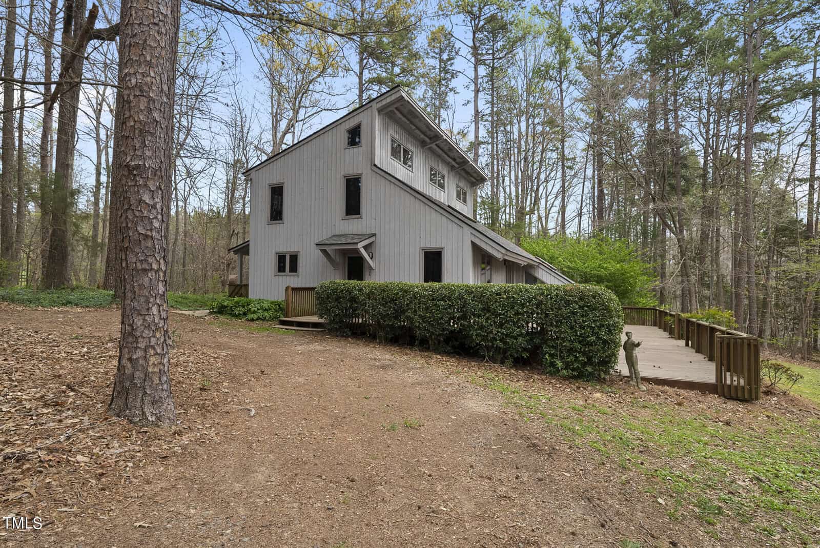 A rustic, multi-story house with a wooden exterior is surrounded by tall trees and greenery. A wooden deck extends from one side, and trimmed bushes line the house. The setting is forested and serene.