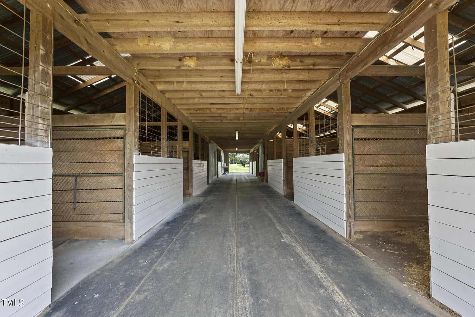 Interior of a long wooden horse stable, with white planks on the lower half of the stalls and open barred sections above. The ceiling is high with exposed beams, and natural light filters in through windows and open doors at the end.