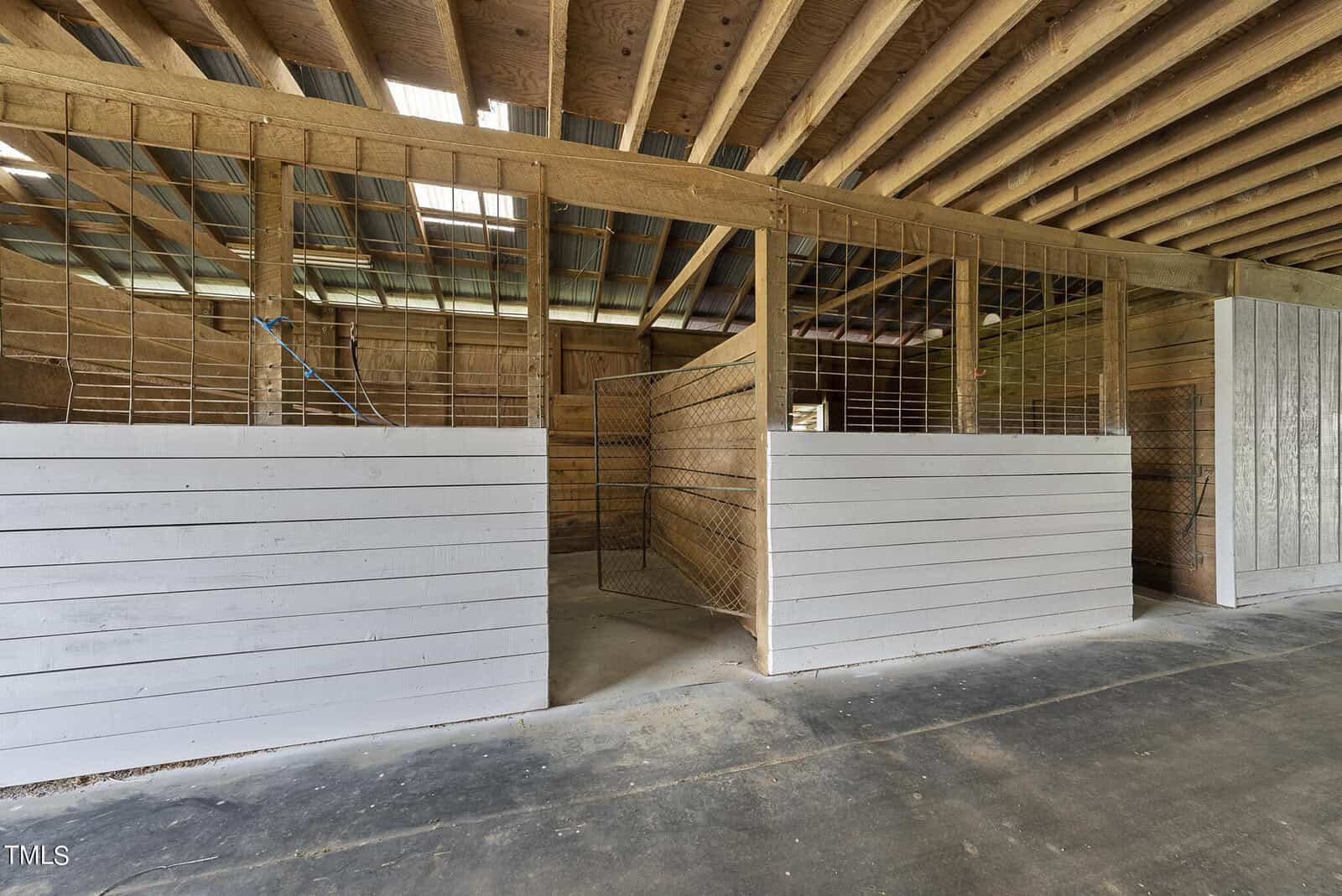 Interior of an empty wooden horse stable with open stalls and a high ceiling. The walls are partially painted white, and the ceiling is constructed with exposed beams. Natural light filters through the windows above.