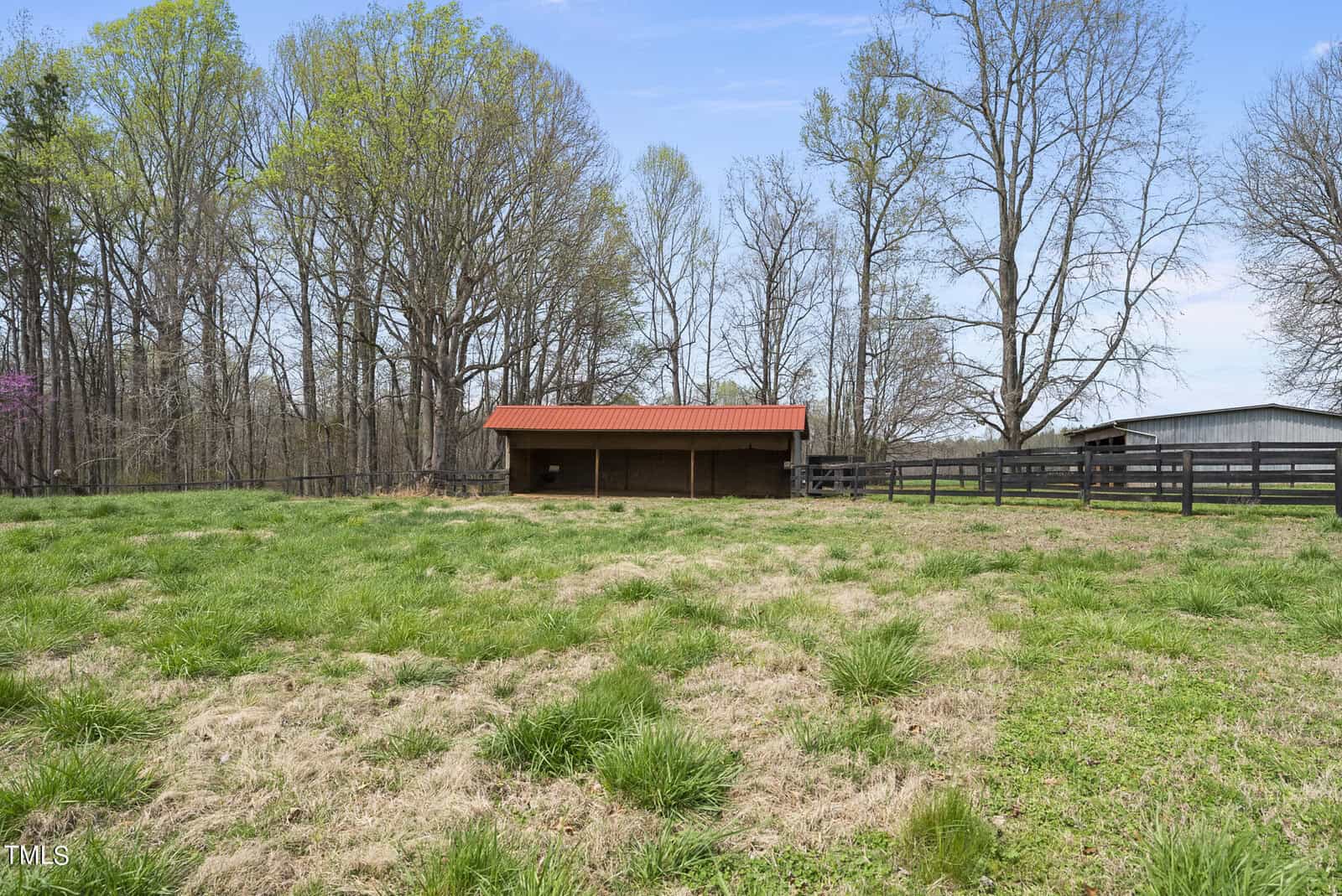A grassy field with a simple wooden shelter topped by a red roof, surrounded by tall trees and a wooden fence, under a clear blue sky.