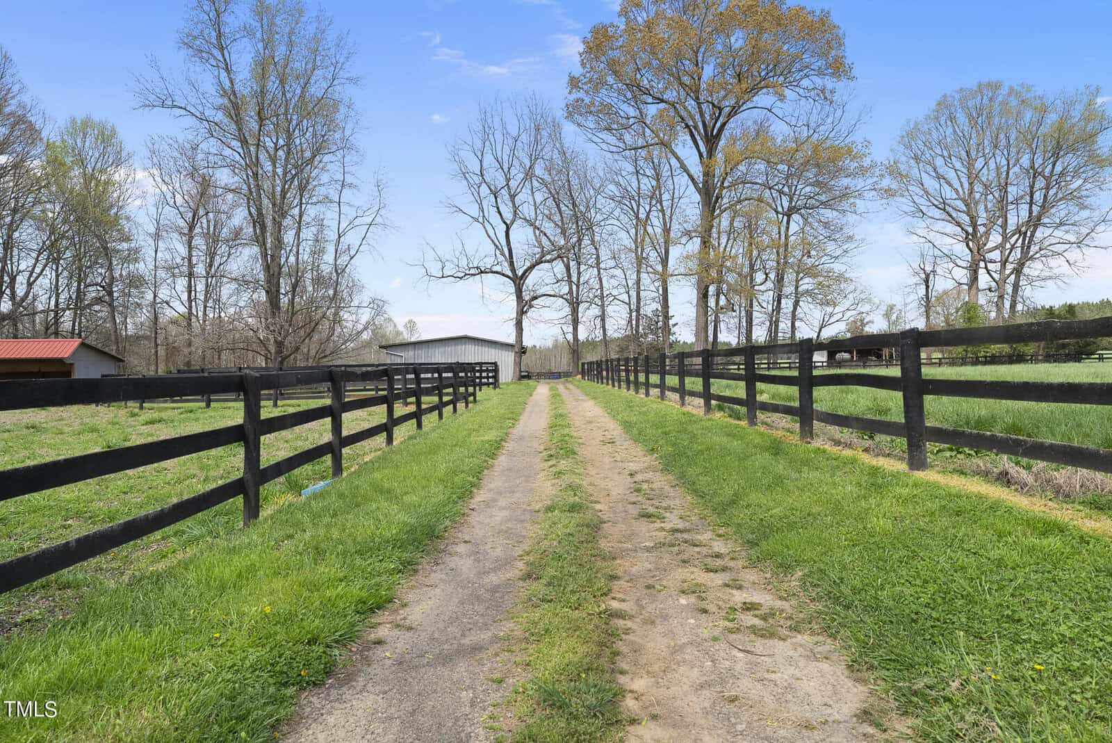 A rural dirt path is shown, flanked by black wooden fences and green grass. Tall, leafless trees line the pathway. A barn-like structure is visible in the distance under a clear blue sky.