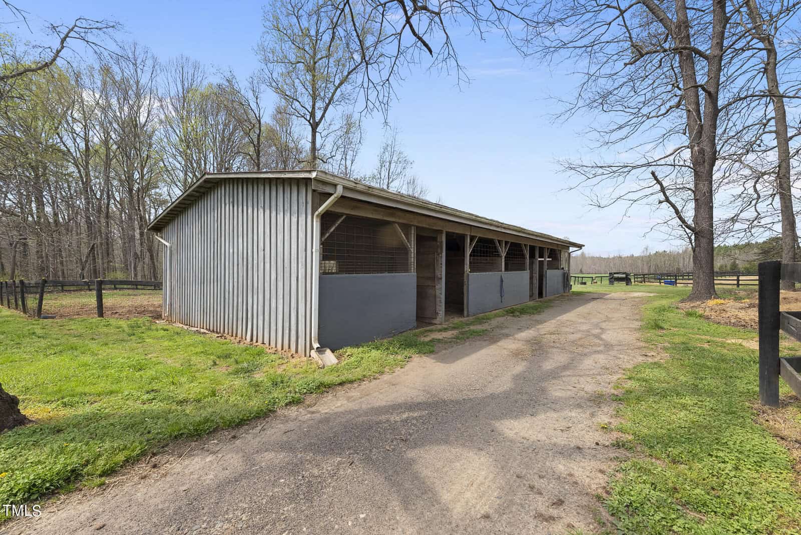 A rustic barn made of gray wood, standing next to a dirt path on a farm. The barn has open stalls, surrounded by leafless trees and a grassy area. A wooden fence lines the surroundings under a blue sky.