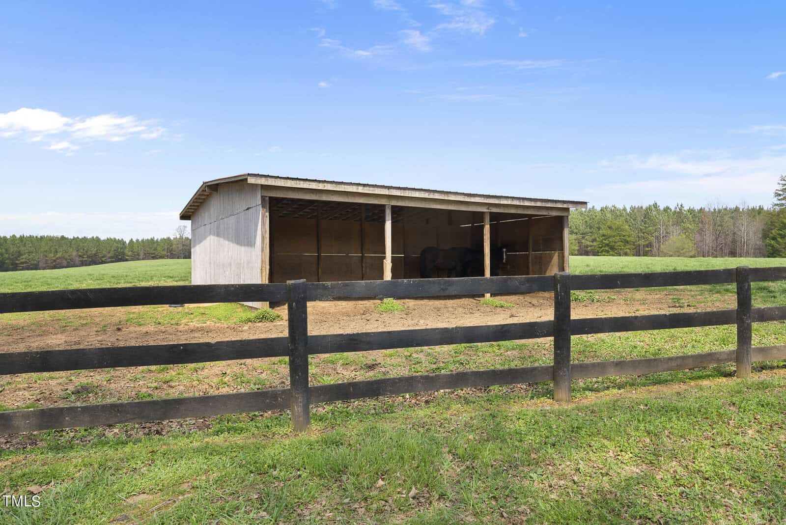 A wooden livestock shelter stands in a grassy field surrounded by a black wooden fence. Trees and a blue sky with clouds are visible in the background.