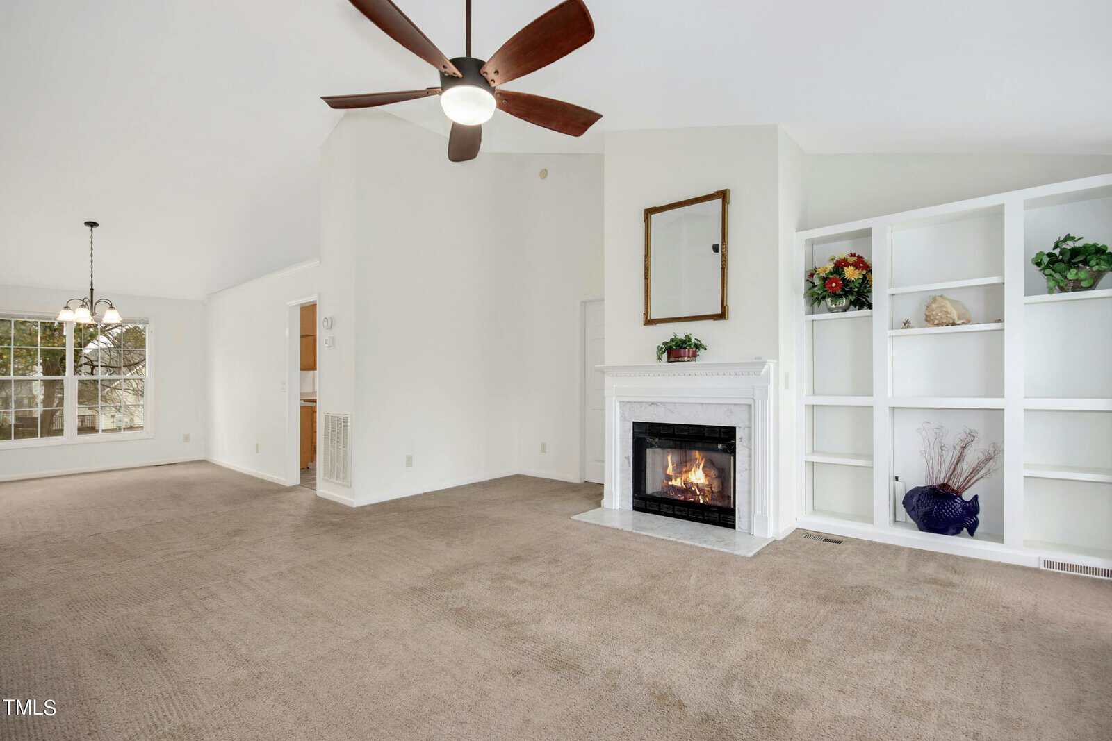 A spacious, carpeted living room with a white fireplace and built-in shelves. A mirror is above the fireplace, and the shelves hold vases and flowers. A ceiling fan is above, and a window provides natural light. An adjoining dining area is visible.