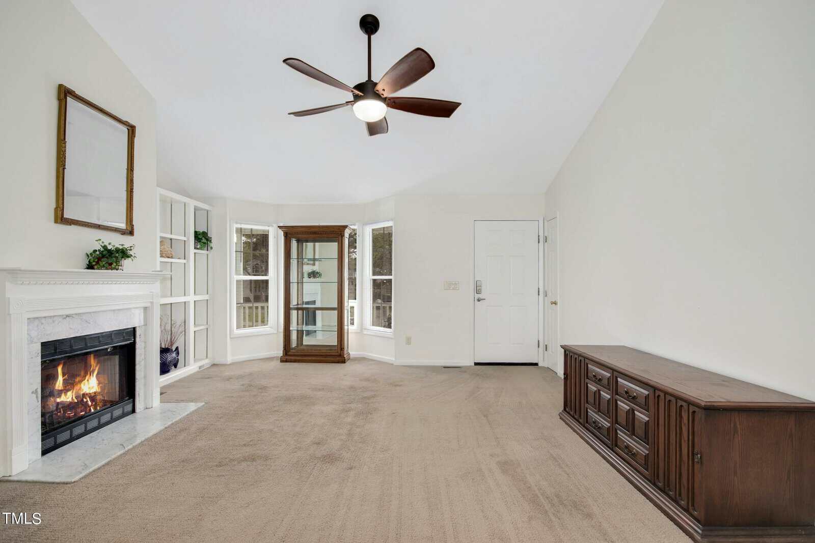 A spacious living room with a ceiling fan and a lit fireplace on the left. Large windows bring in natural light. The room features neutral carpet, a wooden sideboard on the right, and a white paneled wall next to the main door in the background.