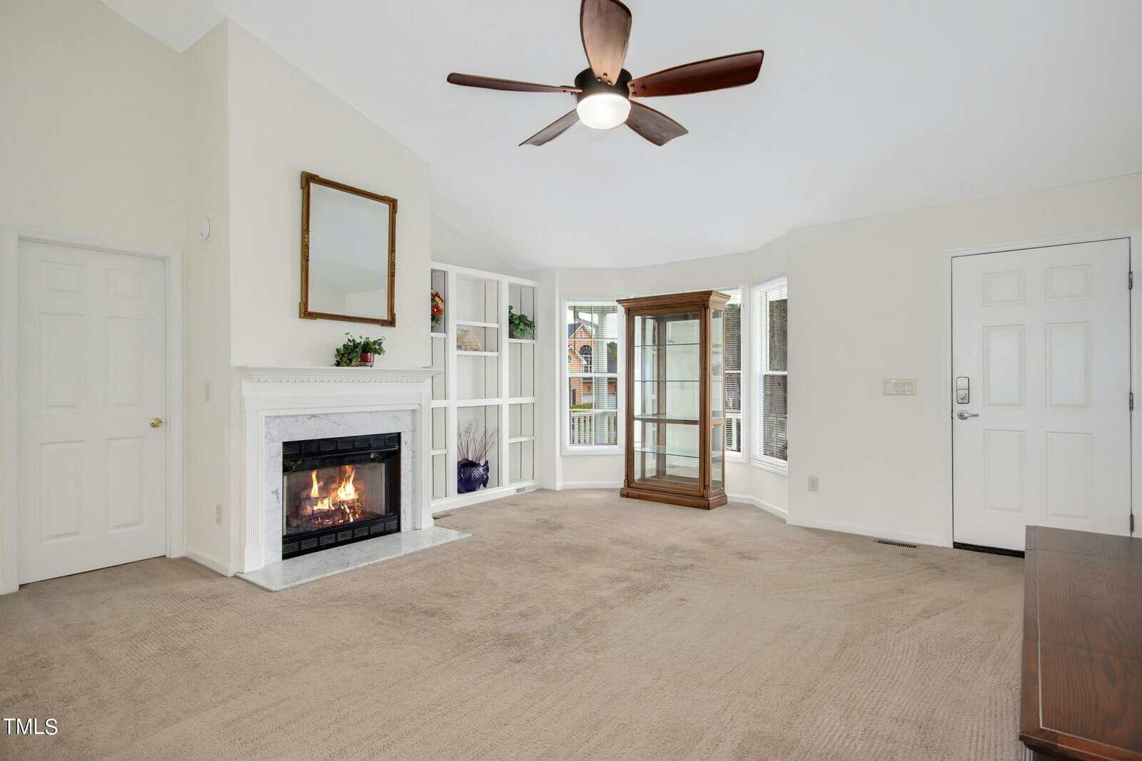A bright living room with a lit fireplace, ceiling fan, and carpeted floor. There's a large window, built-in shelves, a corner display cabinet, and a wall mirror above the fireplace. Two closed doors are visible on the left and right walls.