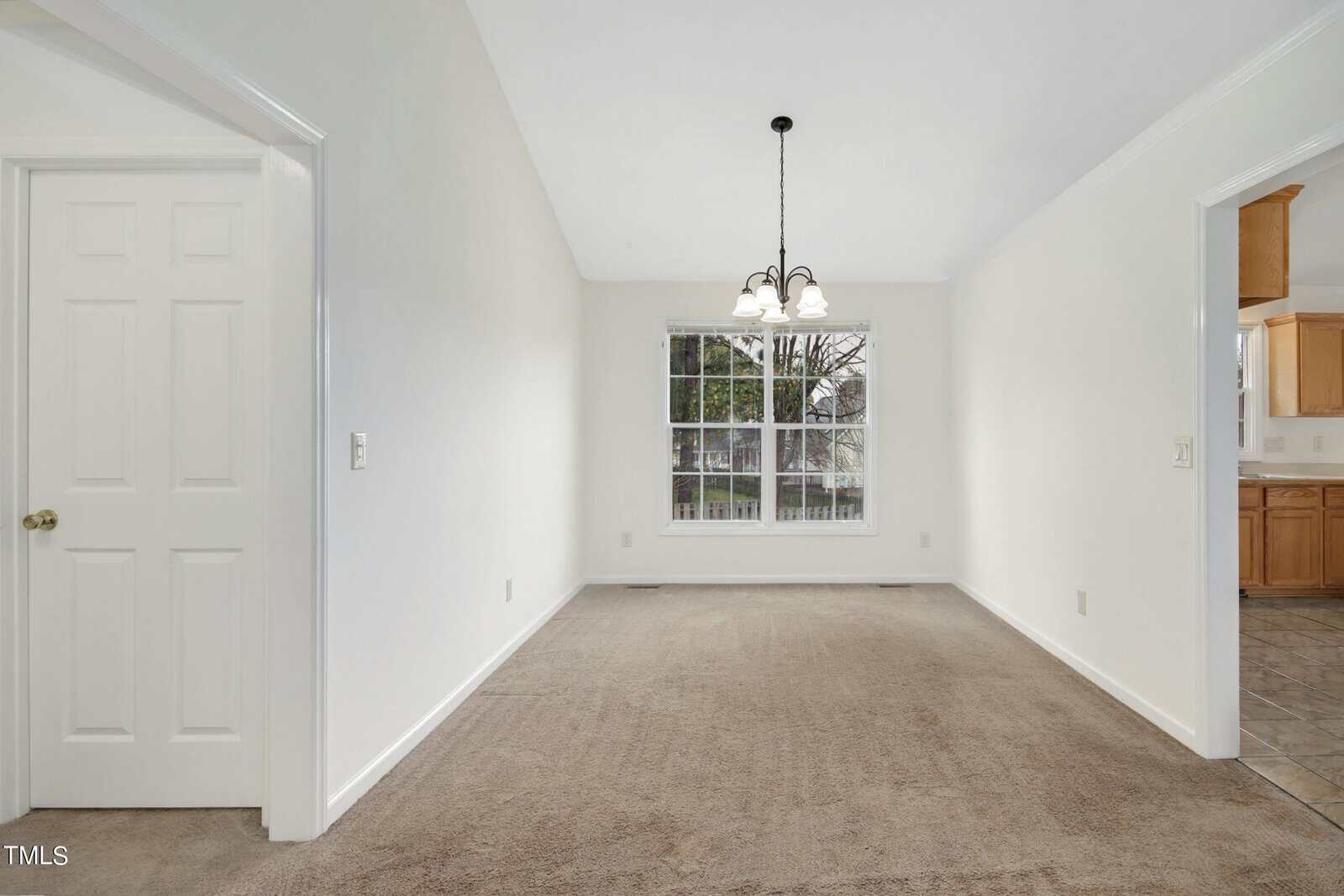 An empty room with white walls, beige carpet, and a large window. A black chandelier-style light fixture hangs from the ceiling. Nearby is an open doorway leading to a tiled kitchen.