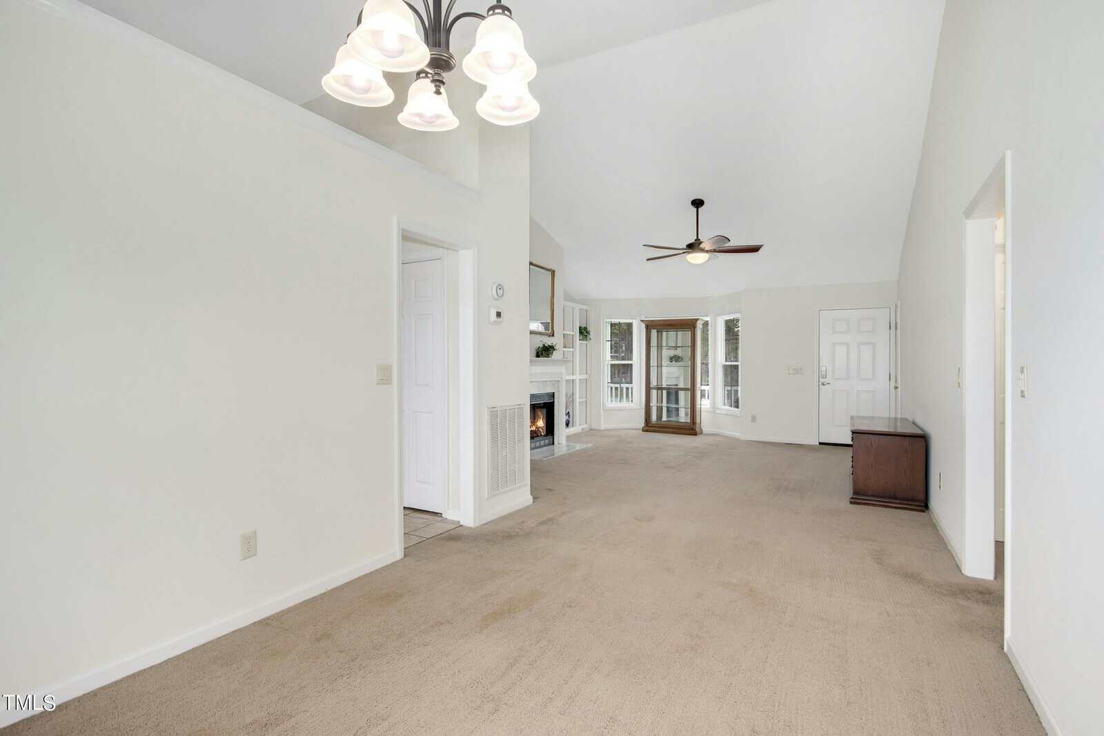 A spacious living room with beige carpet and white walls. It features a ceiling fan, chandelier, and a fireplace with built-in shelves. Sliding glass doors lead to a porch. Multiple doorways open to adjacent rooms. Natural light fills the space.