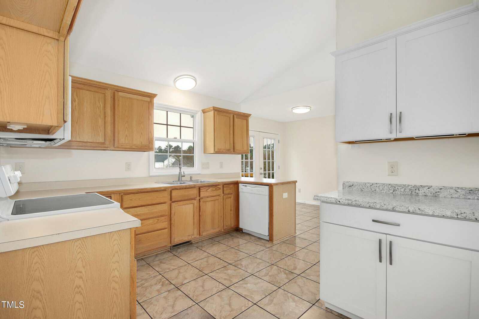 A bright kitchen with wooden cabinets, white countertops, and a tiled floor. There is a white dishwasher beneath the counter, a large window above the sink, and a door leading outside, allowing natural light to fill the space.