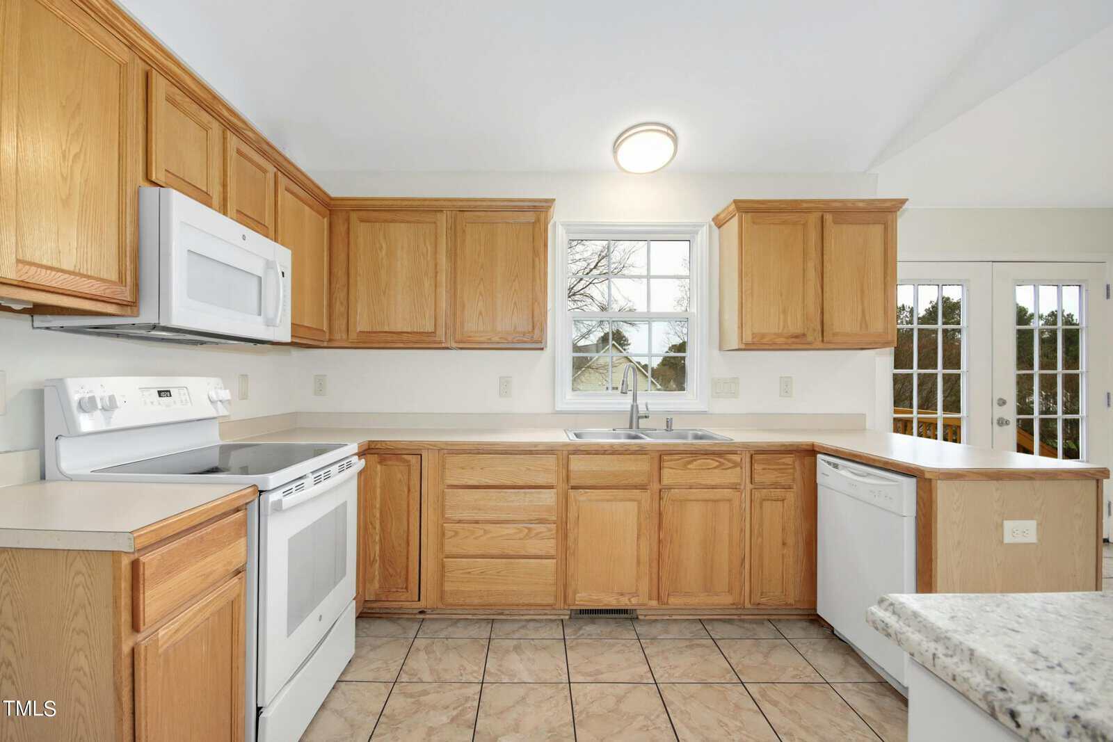 A bright kitchen with wooden cabinets, a white microwave, stove, and dishwasher. There's a large window above the sink and tiled flooring. French doors on the right lead outside, providing ample natural light.