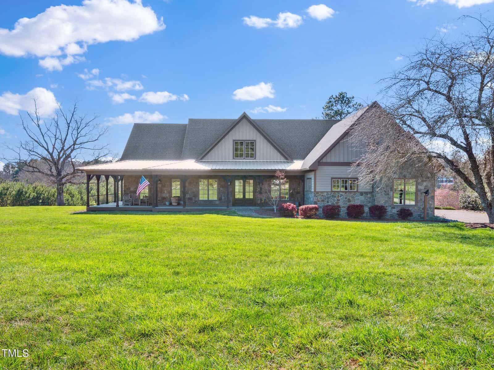 A large house with a wraparound porch and gabled roof sits on a spacious, well-manicured lawn under a blue sky. An American flag hangs by the entrance. Leafless trees are on either side of the lawn.