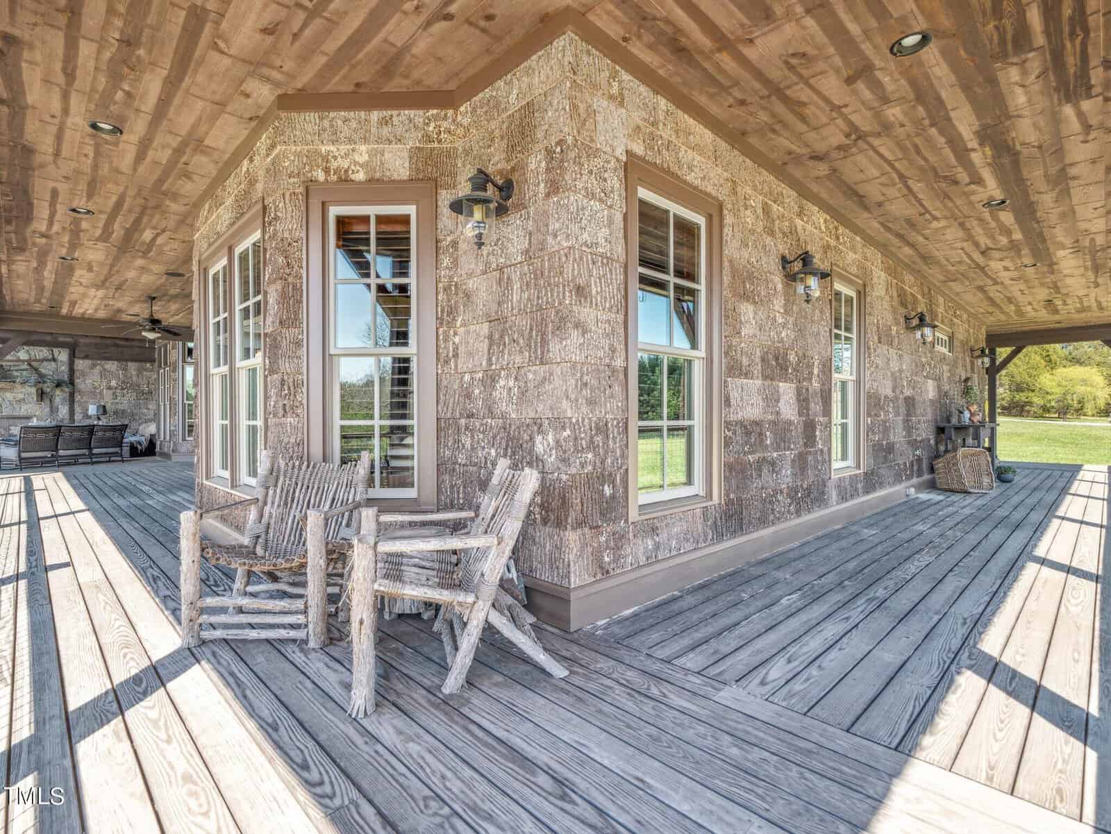 Rustic wooden porch with two weathered chairs and a table, surrounded by a wooden railing. The house exterior features large windows and a log cabin style design. Open green field in the background under a clear blue sky.