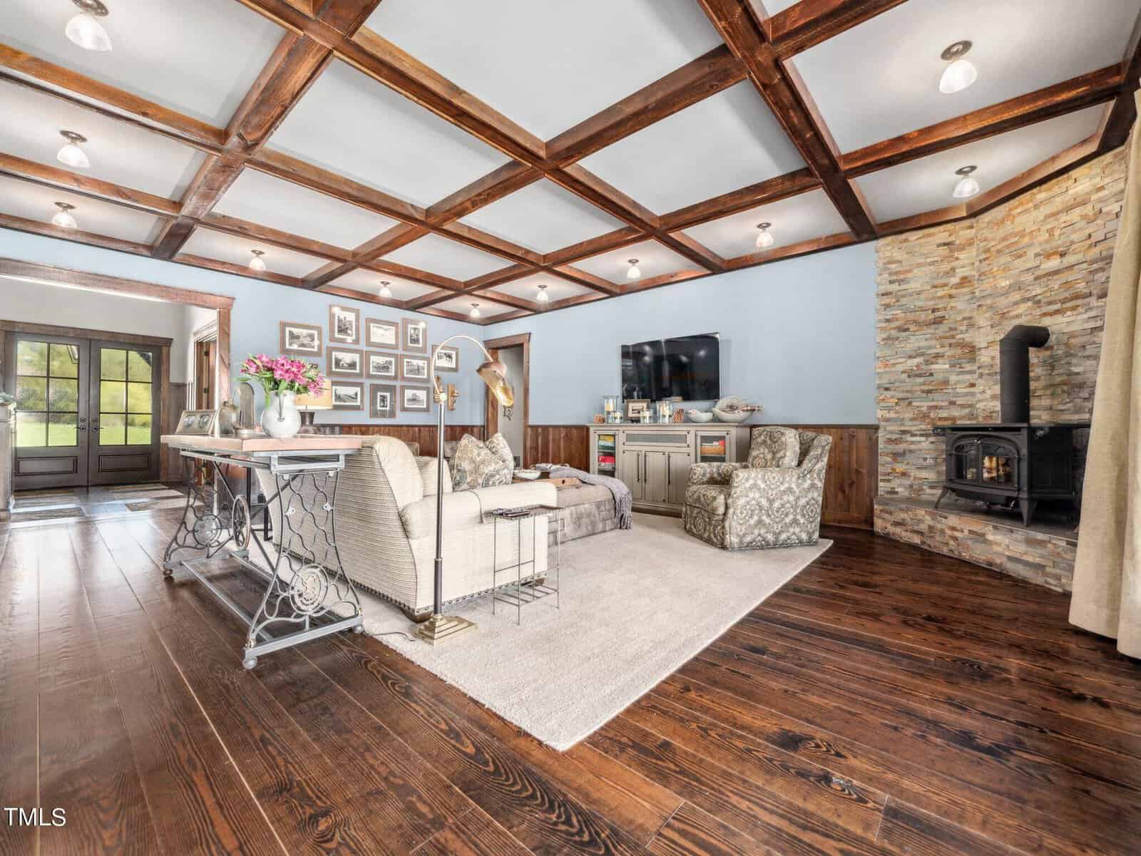 Spacious living room with wooden beam ceiling and hardwood floors. Features a stone fireplace with a black stove, a cozy seating area, a wall-mounted TV, and decorated with framed photos. A large table with flowers is near the entrance.