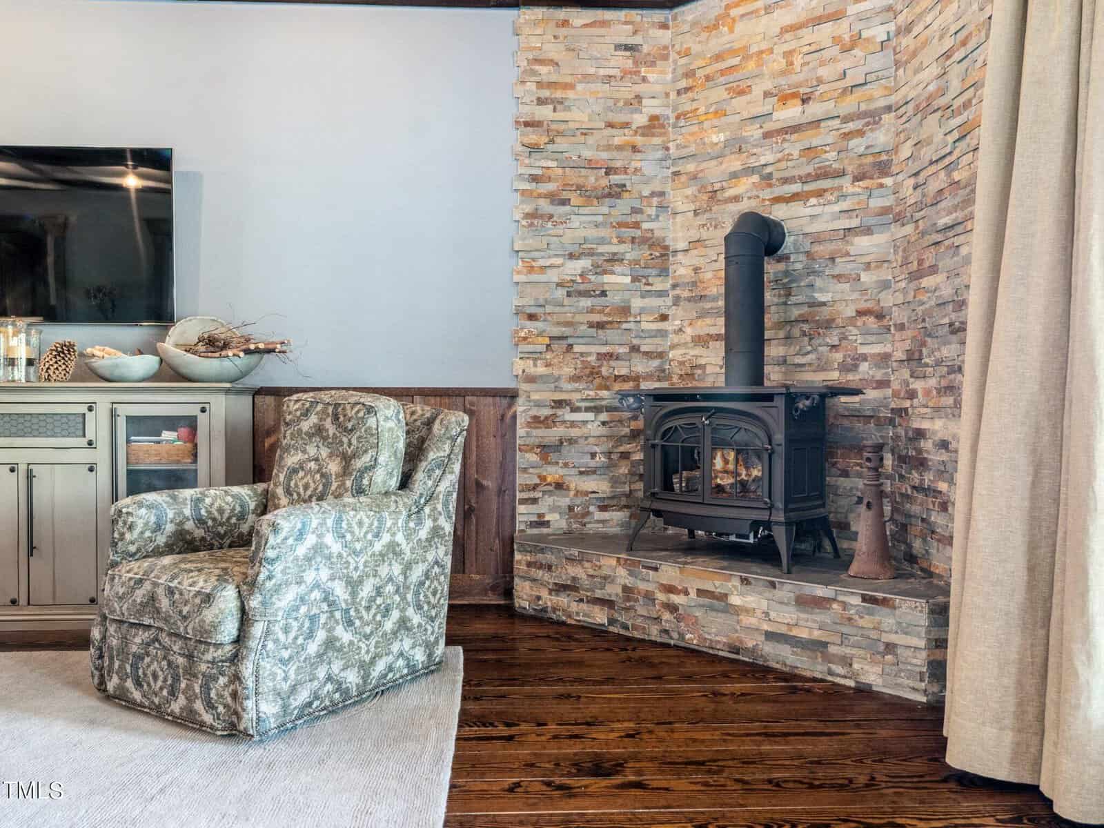 A cozy living room corner featuring a stone fireplace with a black wood-burning stove. A patterned armchair sits on a light rug, adjacent to a wooden cabinet. Neutral tones and rustic decor give the room a warm, inviting feel.