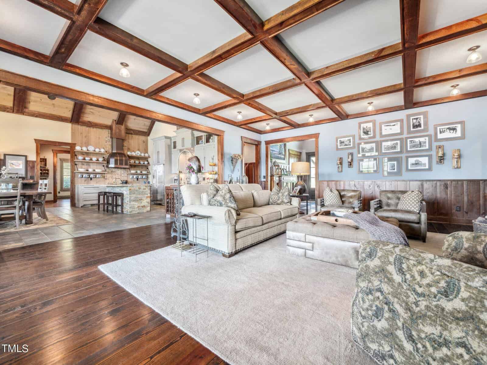 A spacious living room with wooden ceiling beams and hardwood floors. It features two sofas, a cushioned ottoman, and a light-colored rug. Various framed pictures adorn the walls, and the room leads into an adjacent dining area.