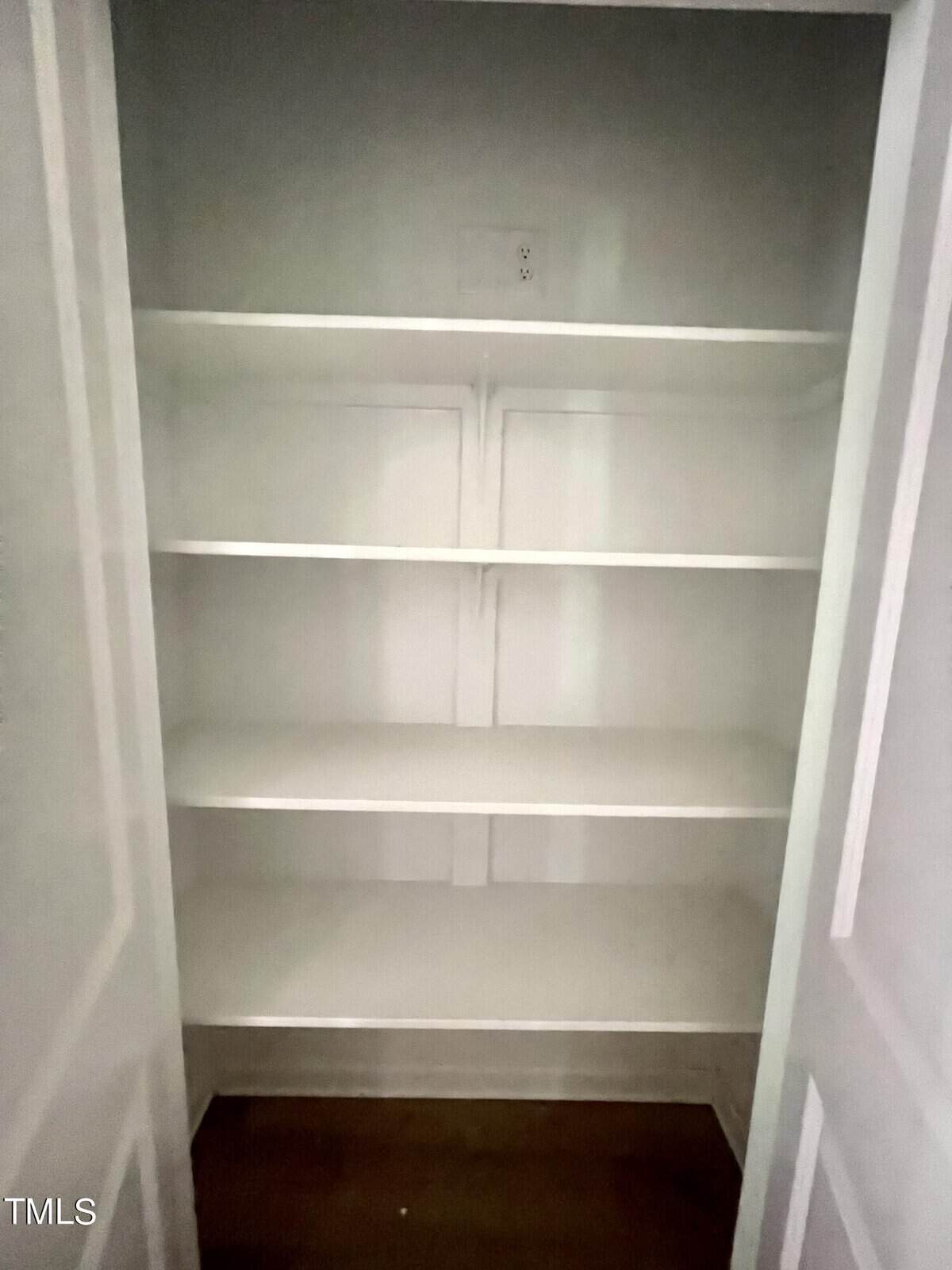 A small, empty white closet with four shelves and a wooden floor, viewed with the doors open. There is an electrical outlet on the back wall above the top shelf.