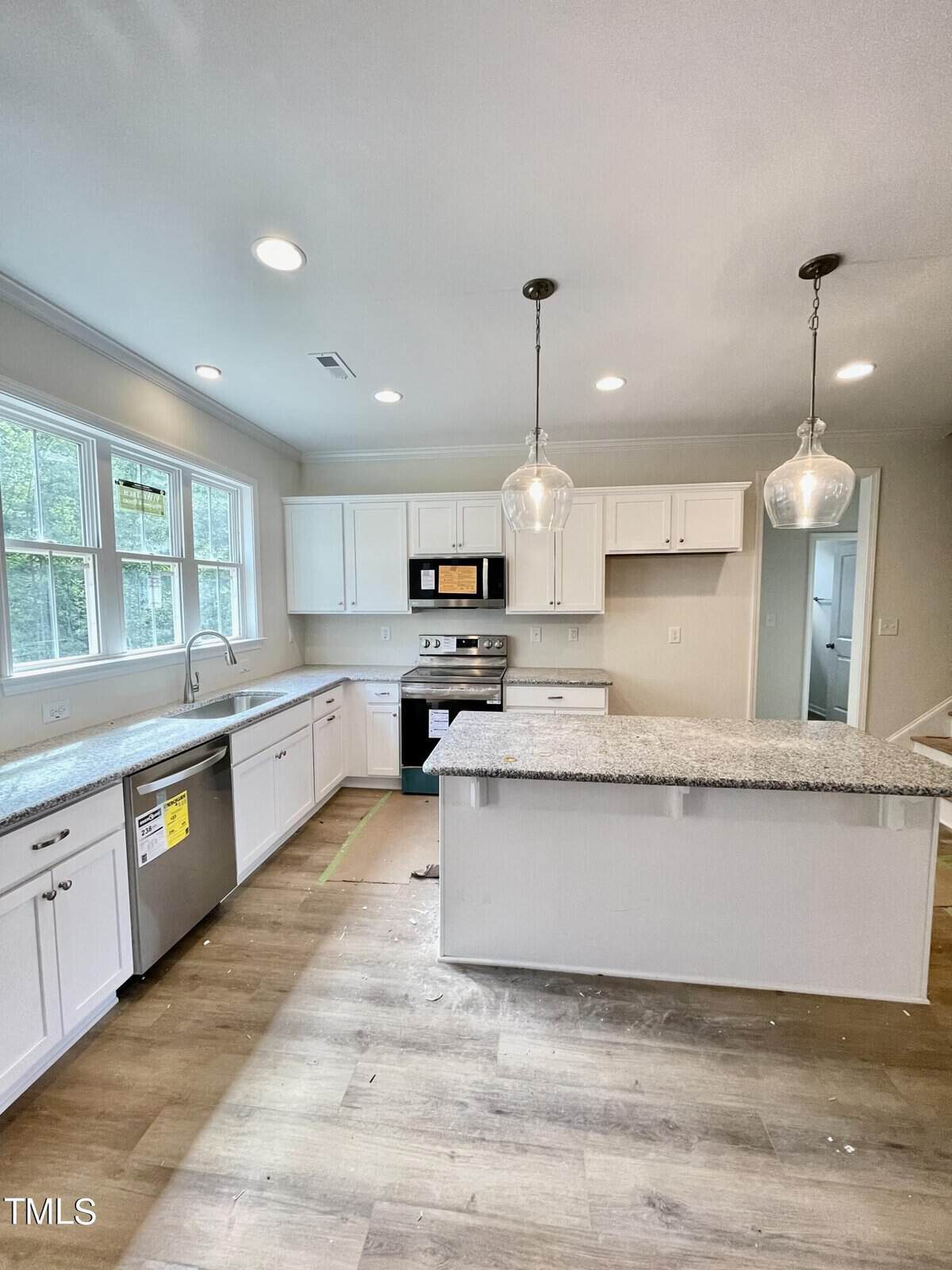 Modern, bright kitchen with white cabinets, stainless steel appliances, granite countertops, a central island with pendant lights, large windows, and wood flooring. Some appliances still have labels on them.