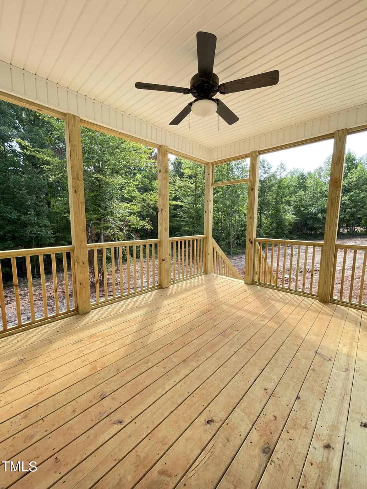 A covered wooden porch with railings and a ceiling fan, overlooking a yard with trees and greenery in the background. The porch has natural wood flooring and posts.