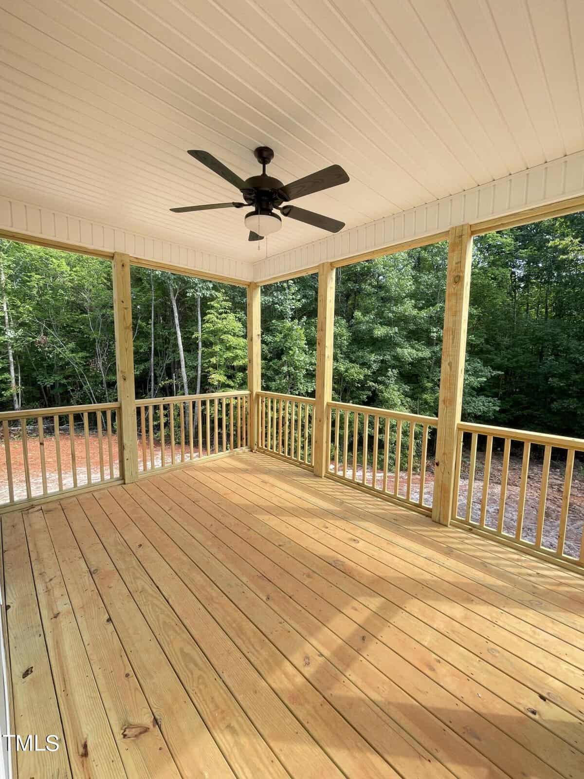A screened-in porch with a wooden floor and railing, surrounded by trees. The ceiling is white with a black ceiling fan in the center, and sunlight is streaming onto the porch.