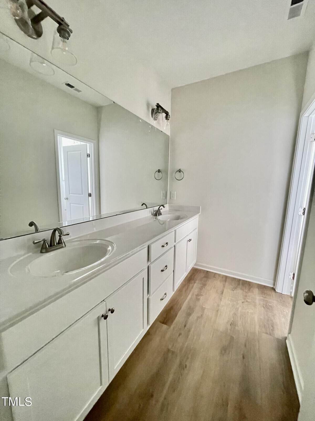 Bright bathroom with a long white vanity, single sink, silver faucet, and large mirror. White cabinets and wood-look flooring. Two doors visible, one open, one closed. Minimalist decor.