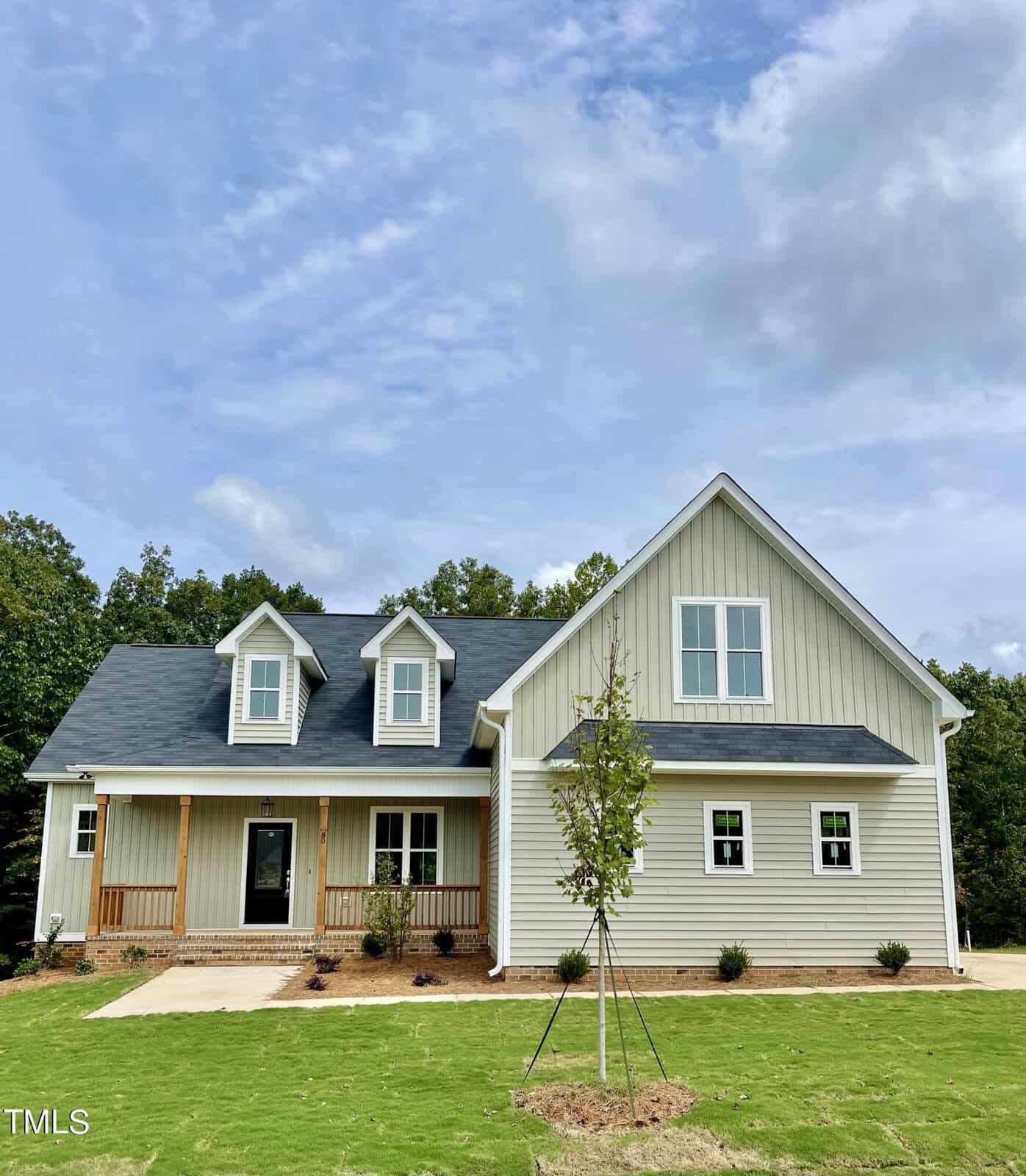 A modern two-story house with gray siding, white trim, dormer windows, and a covered front porch, set on a green lawn with young trees and surrounded by trees under a partly cloudy sky.