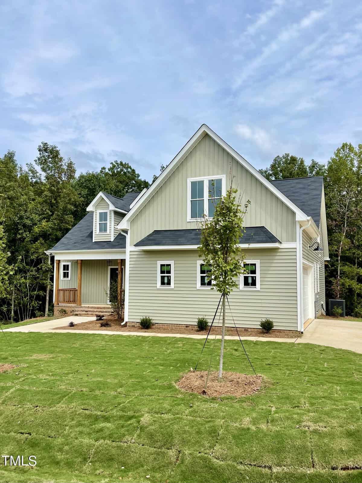 A modern light gray house with white trim sits on a freshly sodded lawn. The home features a covered front porch with wood accents and is surrounded by trees under a blue sky.