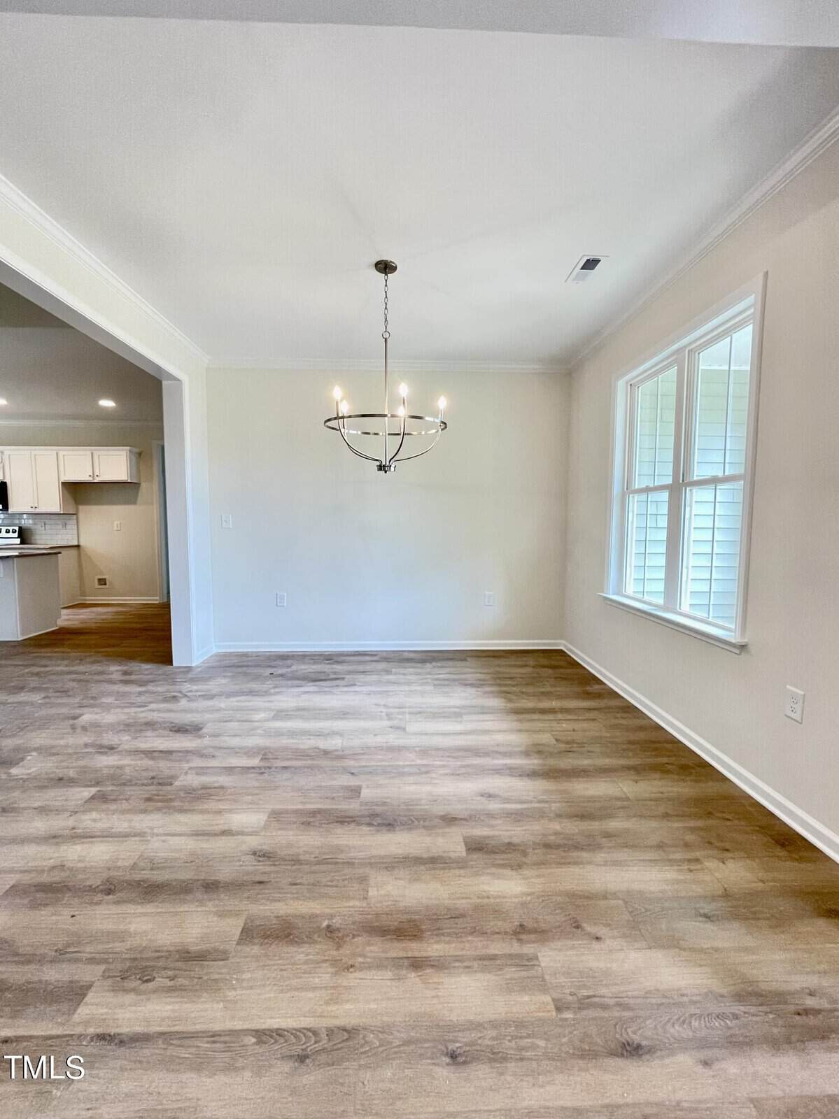 A bright, empty dining room with light wood flooring, a modern chandelier, large window, and an open doorway leading to a white kitchen.