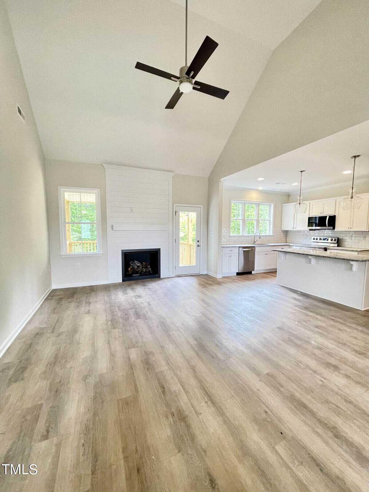 Open-concept living area with light wood floors, a ceiling fan, fireplace, large windows, and a modern kitchen with white cabinets, stainless steel appliances, and pendant lighting. Neutral walls and vaulted ceiling.