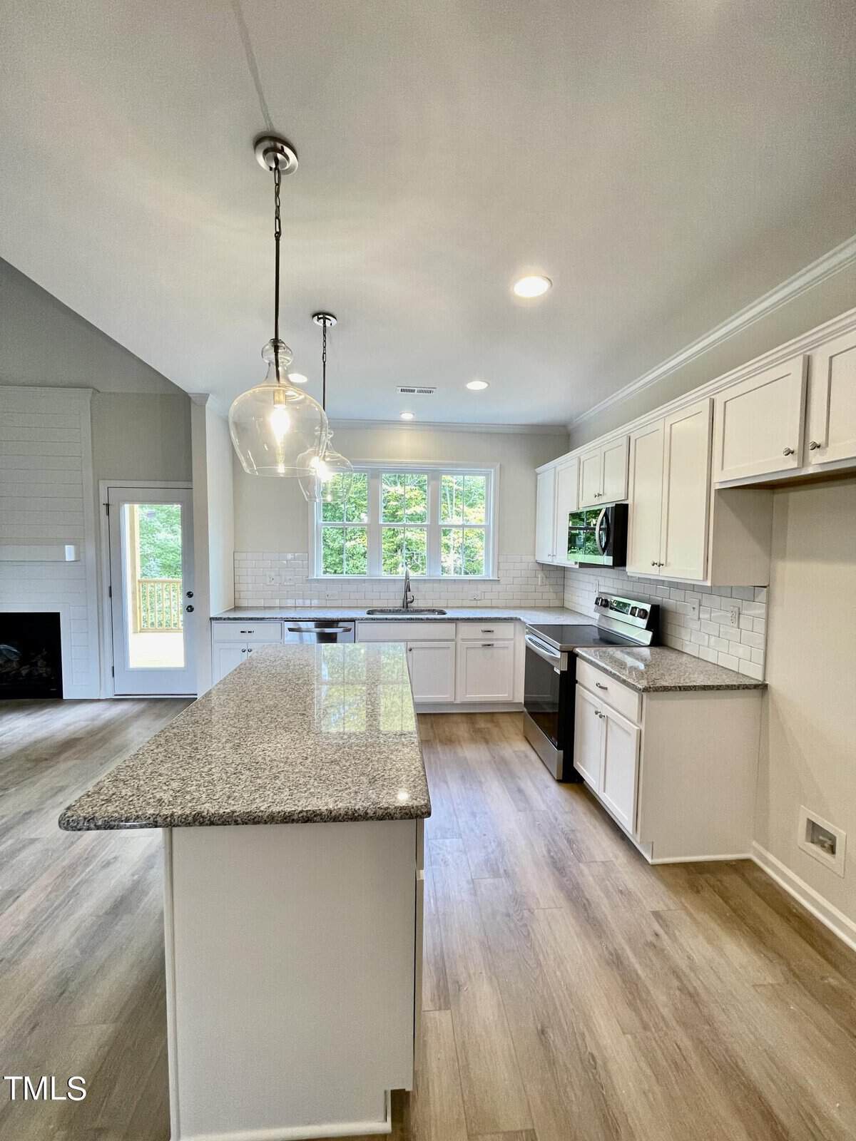 Modern kitchen with white cabinets, granite countertops, stainless steel appliances, a large island, pendant lights, and wood floors. Sunlight streams through three large windows above the sink, brightening the open space.