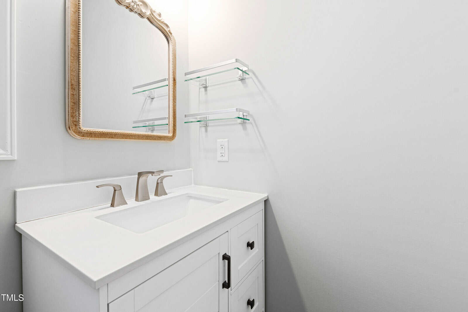 A small bathroom features a white vanity with a modern sink and silver faucet, a gold-framed mirror above, and two glass shelves on the wall. The walls are painted white, creating a clean, minimalist appearance.