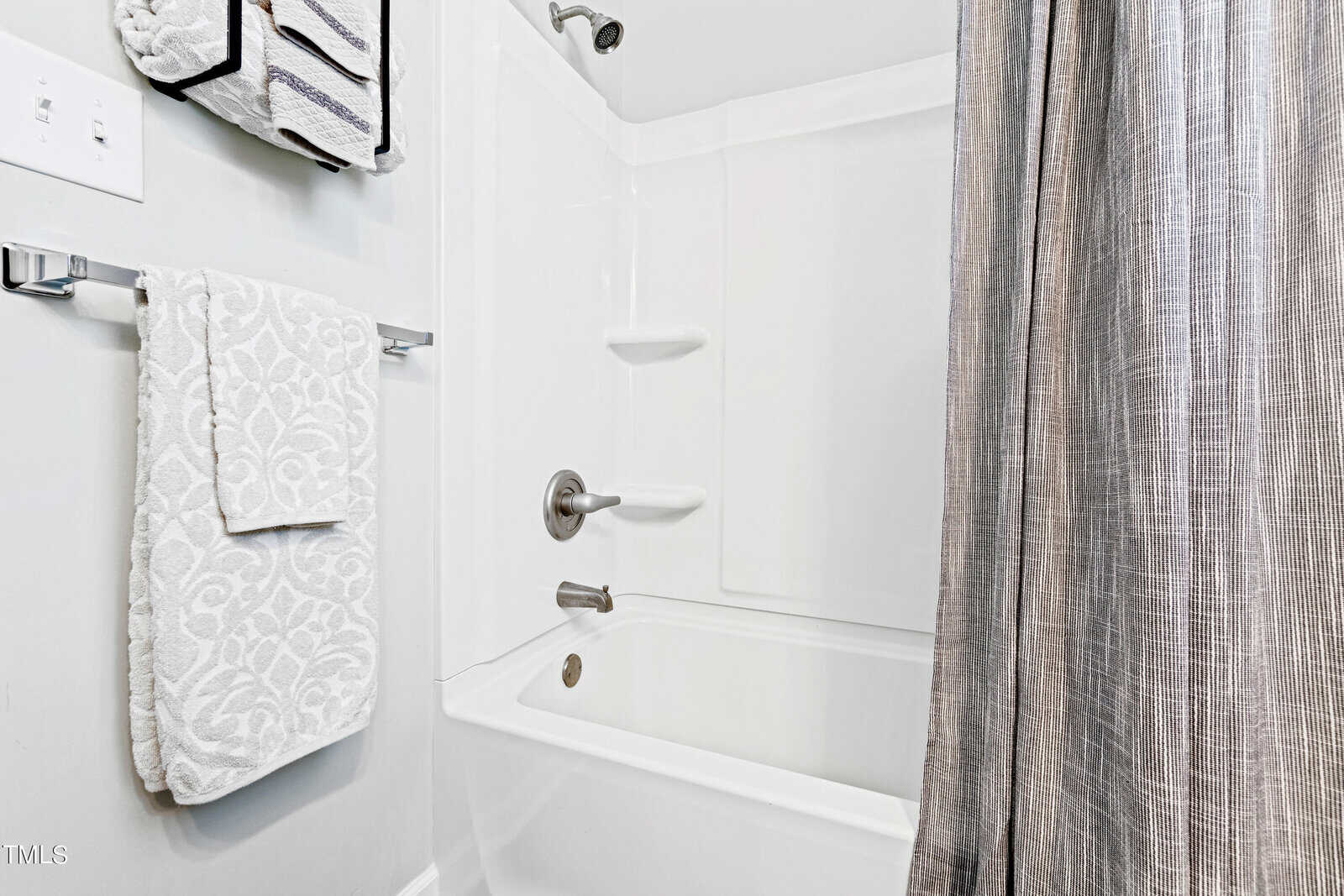 White bathroom with a bathtub and shower combination. A gray shower curtain is situated to the right. Towels are neatly hung on a rack on the wall, with a patterned towel in focus. The room features a simple, clean design.