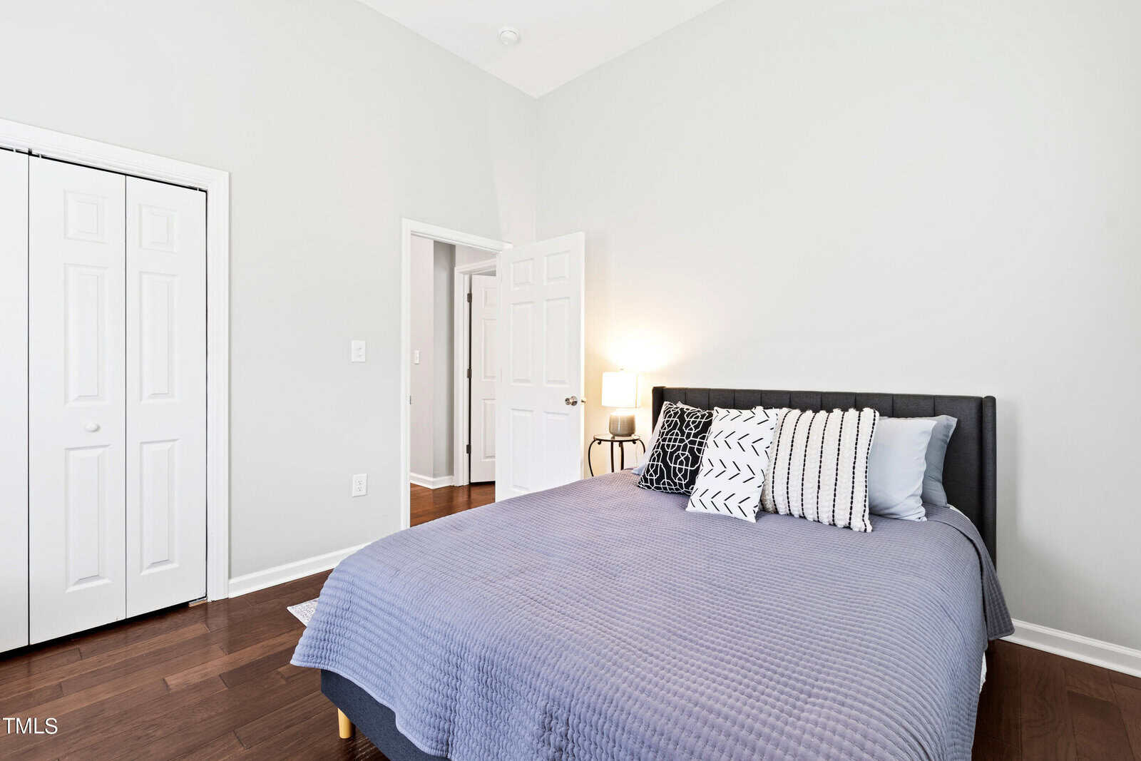 A bright bedroom with a neatly made bed featuring a gray quilt and decorative pillows. The room has white walls, a closed closet with double doors, and a small lamp on a bedside table. The floor is dark hardwood.