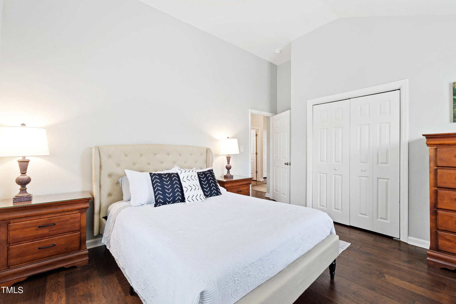 A cozy bedroom with a large bed featuring white and dark blue pillows and a light-colored quilt. Two wooden nightstands with lamps are on either side. A wooden dresser is against the wall, and a closed white door is visible. The room has a soothing ambiance.