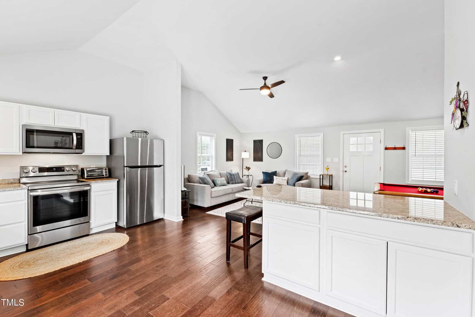 Spacious open-plan kitchen and living room with white cabinets, stainless steel appliances, and a granite countertop. The living area features a sectional sofa and a ceiling fan. A pool table is visible near the brightly lit entrance.