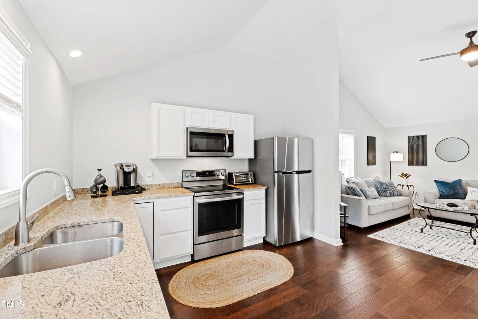 A modern kitchen with white cabinets, stainless steel appliances, and granite countertops. A rug lies on the dark wooden floor. The kitchen opens into a cozy living area with a light-colored sofa, cushions, and a ceiling fan. Natural light fills the room.