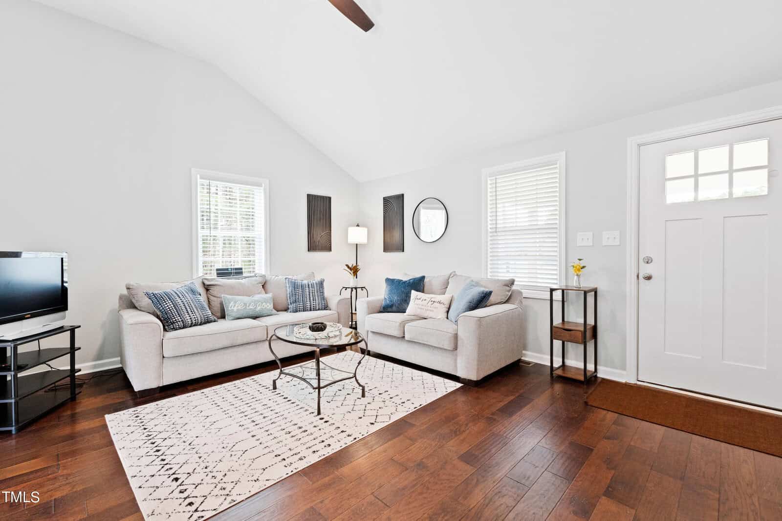 A bright living room with two gray sofas, a round glass coffee table, a white patterned rug, and a TV on a stand. The room features hardwood floors, a white door with windows, and wall decor including a mirror and a lamp.
