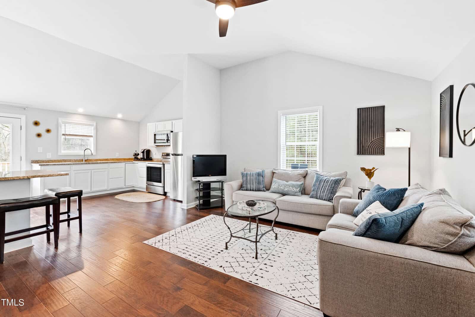 A modern living room with hardwood floors, two gray sofas with blue and white pillows, a glass coffee table on a patterned rug, a small TV, and a floor lamp. The open kitchen has white cabinets, a counter with stools, and stainless steel appliances.