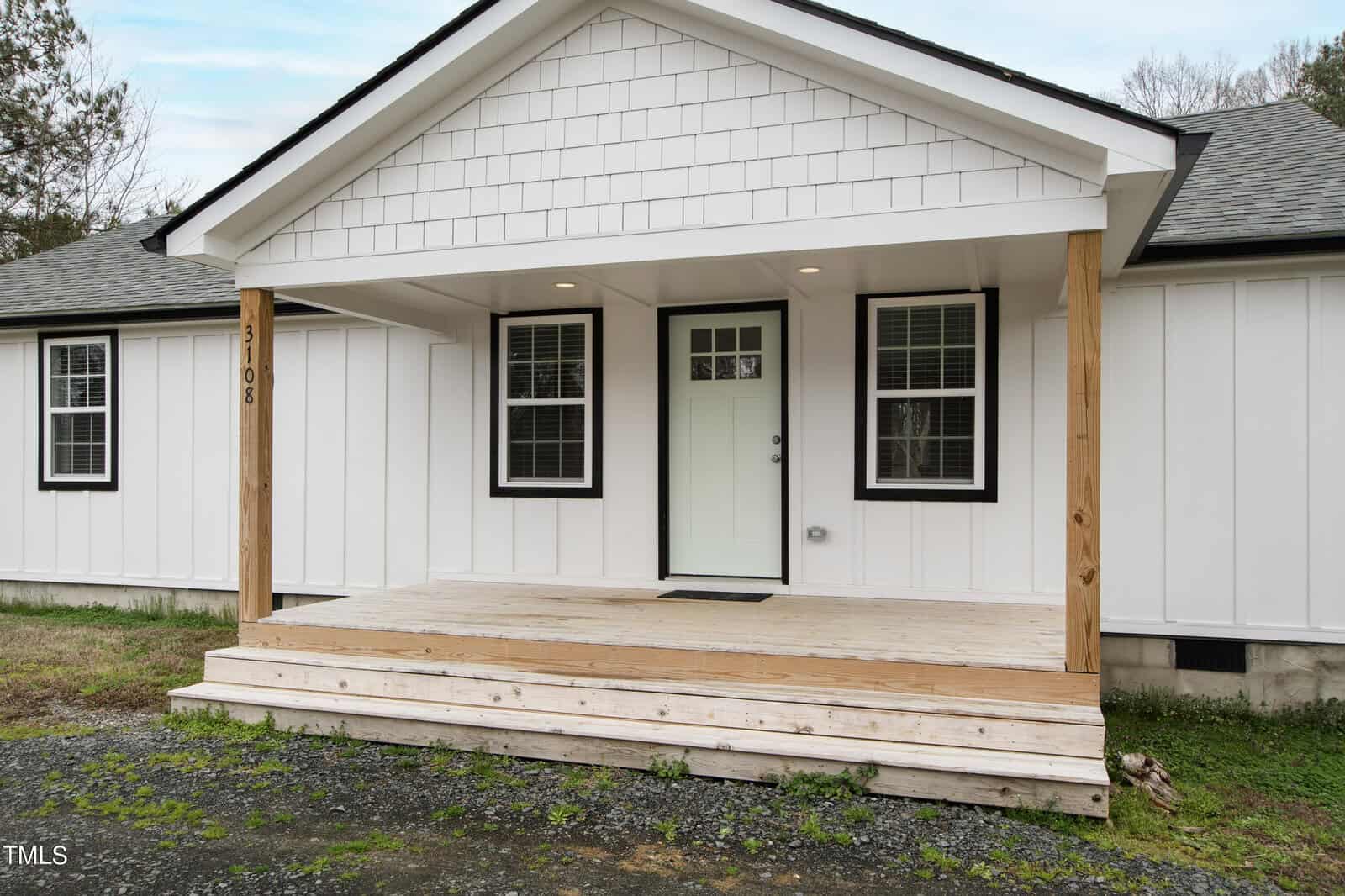 A small, modern white house with a light green front door, black trim, and a wooden porch. The roof is dark gray, and the yard is mostly gravel and grass. The front has a few steps leading up to the door.