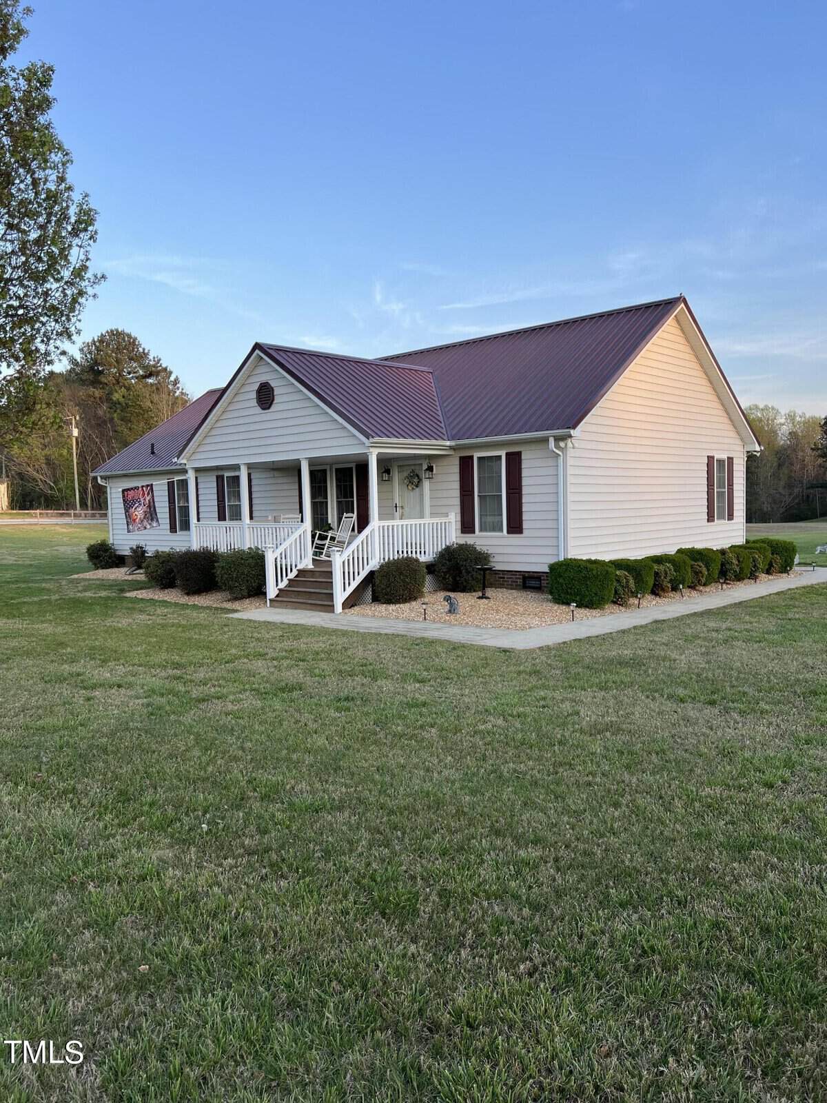 A single-story house with a red metal roof and beige siding, featuring a covered front porch with white railing, surrounded by a manicured lawn and a few shrubs under a clear sky at dusk.